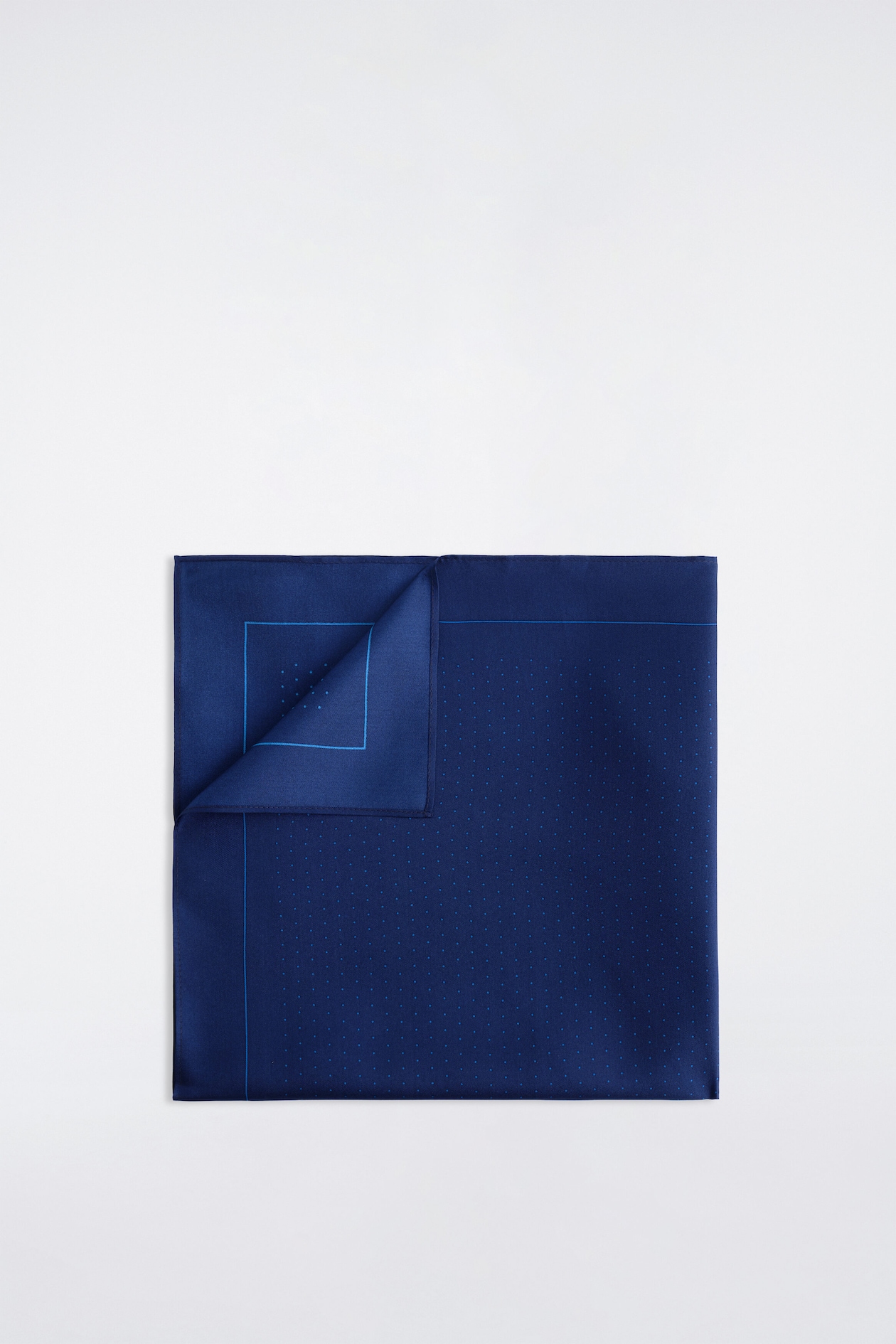 A dark blue silk pocket square is laid flat and facing forward, with one corner folded back to show the reverse side. The pocket square features a pattern of tiny light blue polka dots, framed by a matching thin blue inner border and finished with stitched edges.