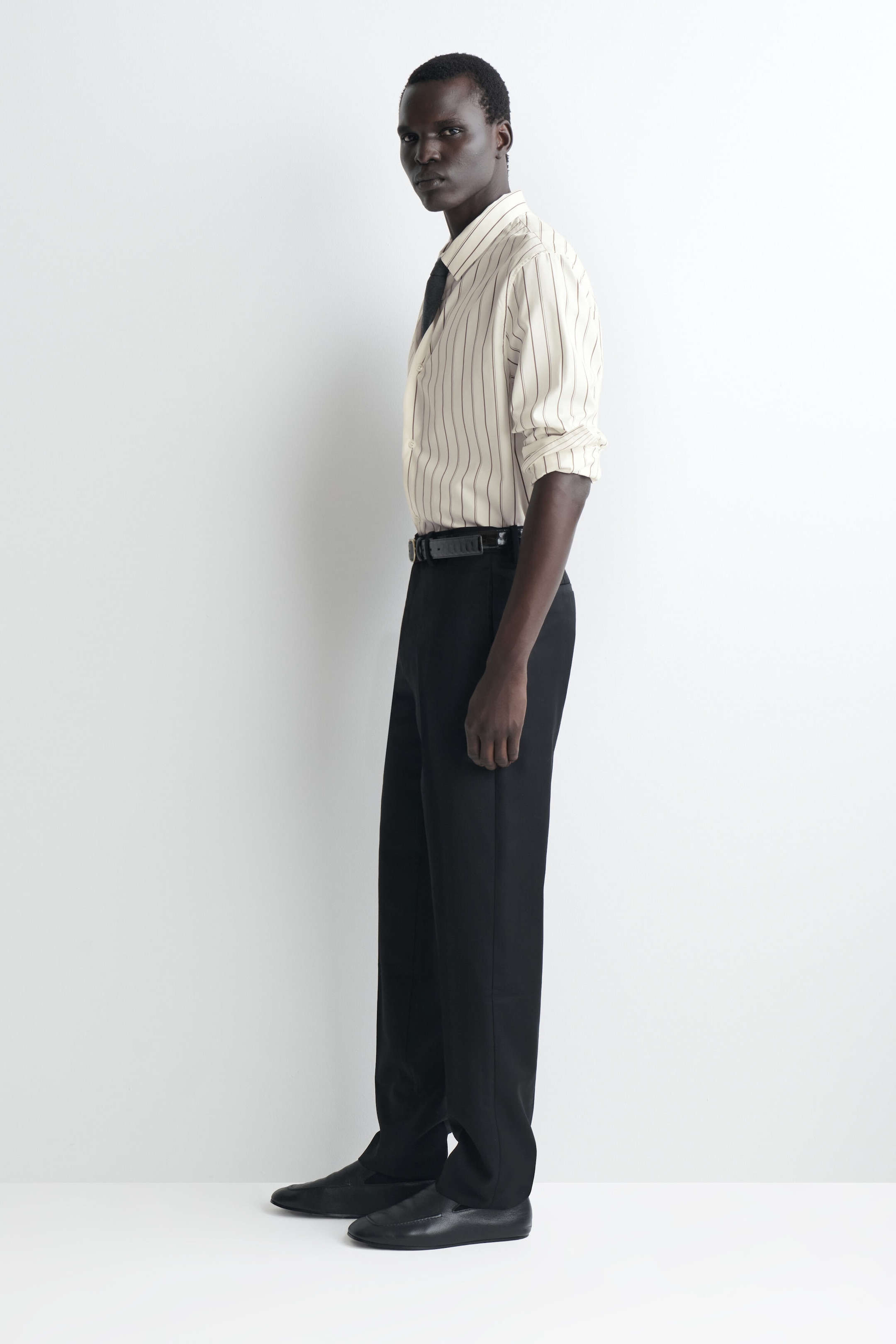These black straight-leg trousers are shown in a profile view facing left, showcasing a regular fit that falls straight to the ankles. They are being styled with a tucked-in striped button-down shirt, a black leather belt, and black loafers. These black straight-leg trousers are shown in a profile view facing left, showcasing a regular fit that falls straight to the ankles. They are being styled with a tucked-in striped button-down shirt, a black leather belt, and black loafers.