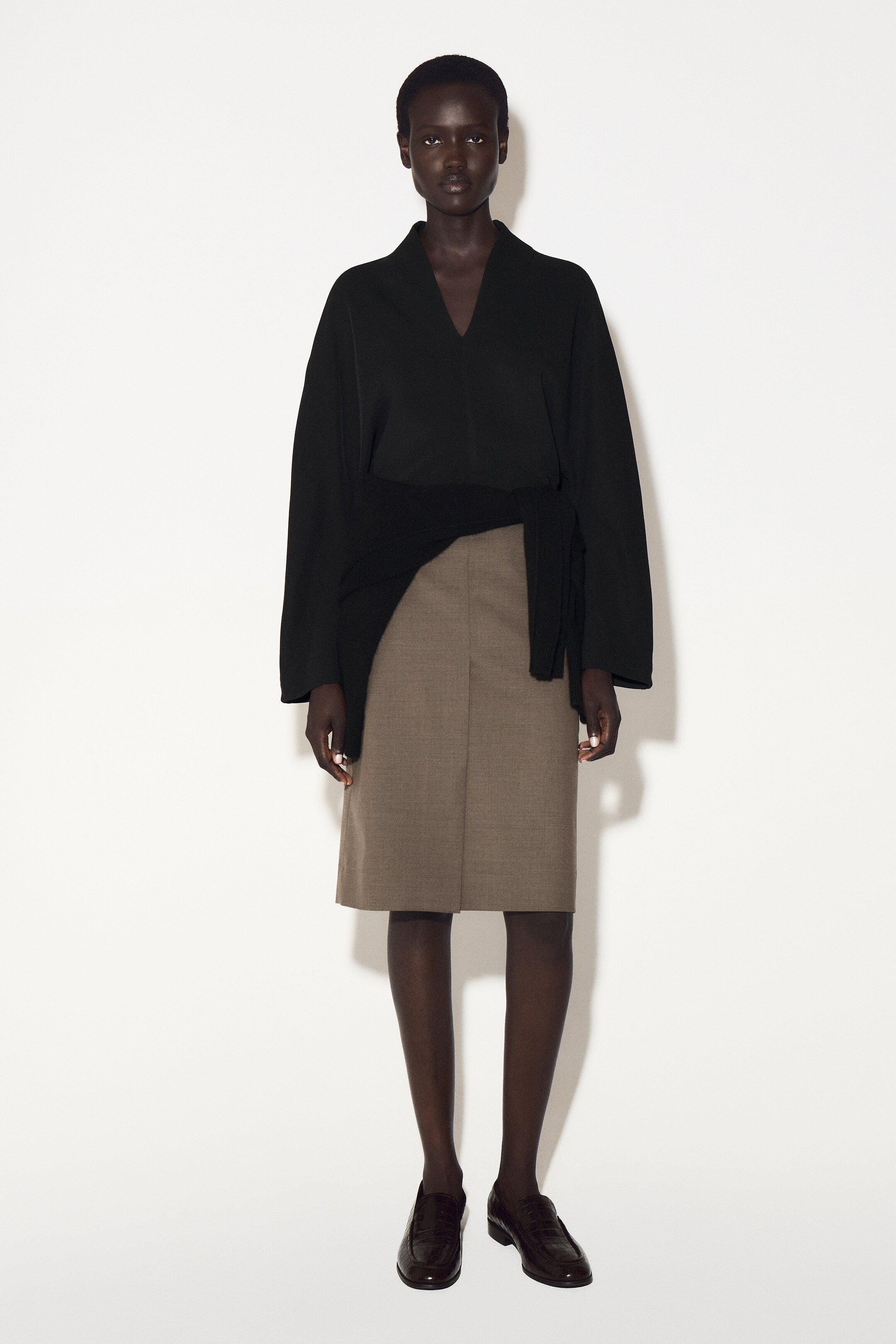 Größeres Bild anzeigen: Eine schwarze Bluse wird frontal präsentiert, mit einem skulpturalen V-Ausschnitt und einem drapierten Kragen. Sie zeigt eine entspannte Passform mit weiten Ärmeln und einem sichtbaren Selbstbindedetail, das die Taille betont und eine blusige Silhouette schafft.