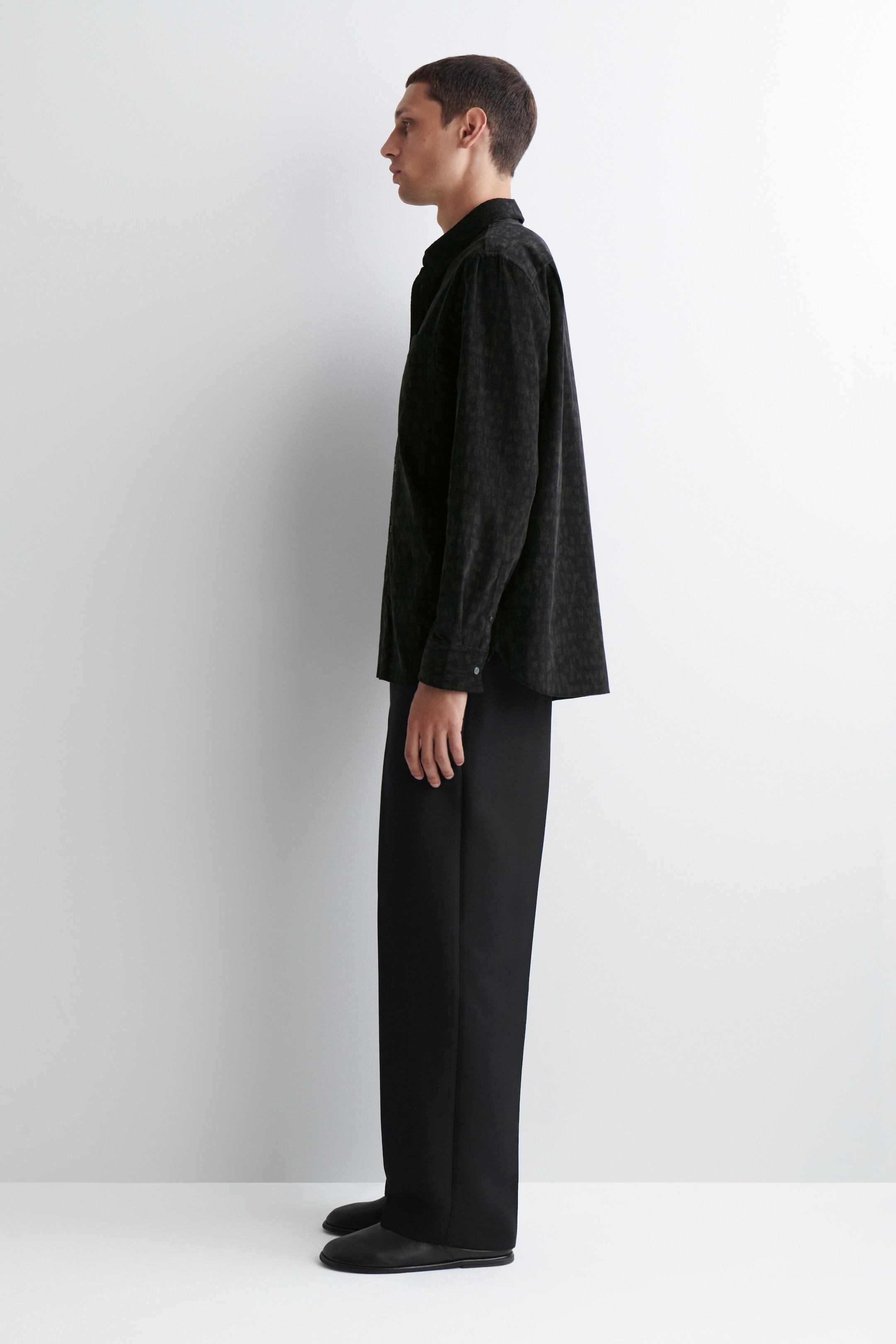 Agrandir l'image: Une chemise en velours côtelé de couleur sombre et à coupe décontractée est présentée sur une personne tournée vers la gauche, avec des manches longues dotées de poignets boutonnés et un col standard.