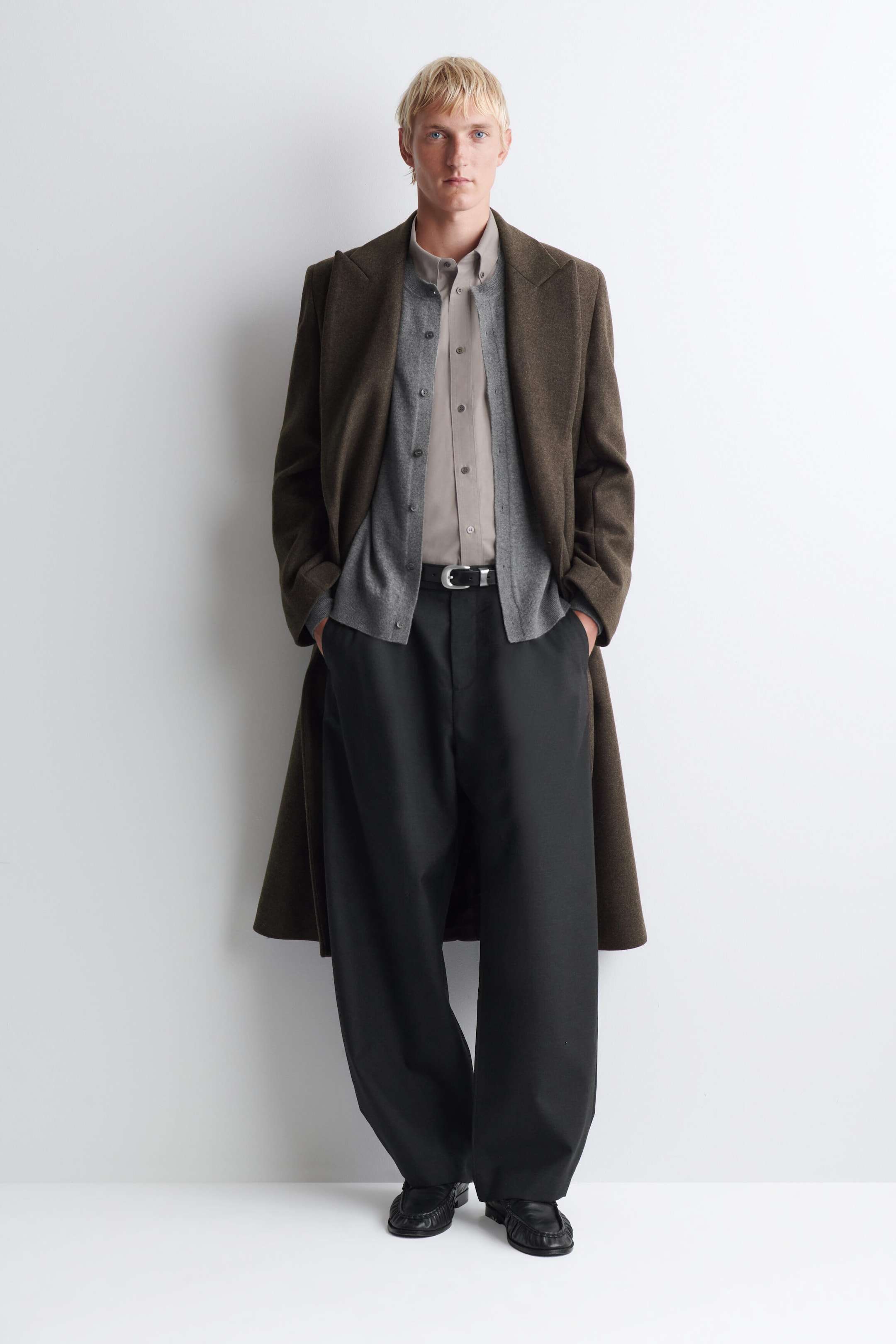 The button-down collar brushed oxford shirt in light taupe is worn facing front, tucked into black trousers with a belt. It is styled layered under a grey button-front cardigan and a long brown overcoat, showcasing its structured collar and buttoned placket.