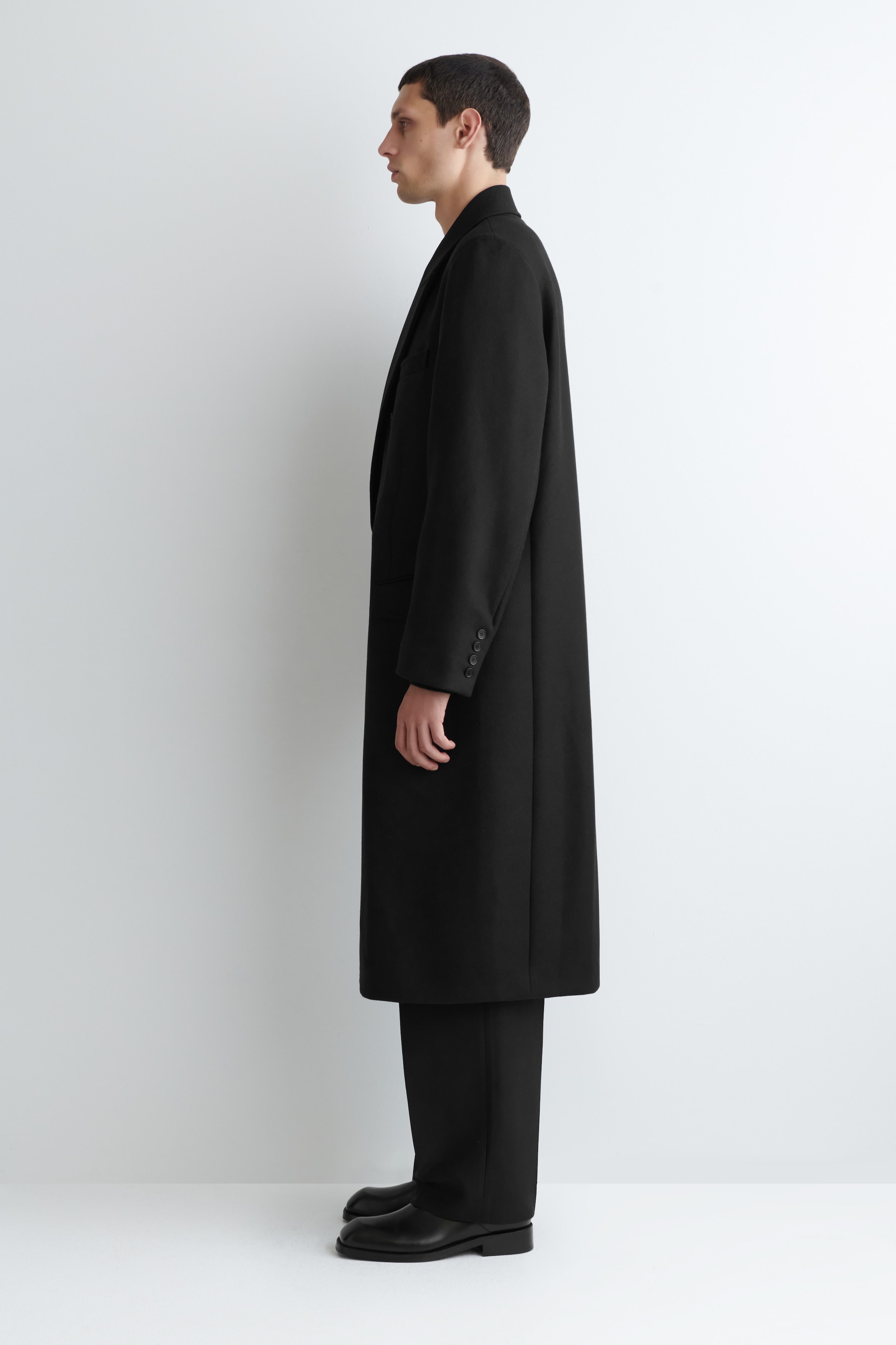 Visualizza immagine più grande: Un lungo cappotto nero è presentato in profilo laterale, con revers a dente e quattro bottoni sul polsino della manica destra, che si estende sotto il ginocchio e si sovrappone a pantaloni e scarpe nere.