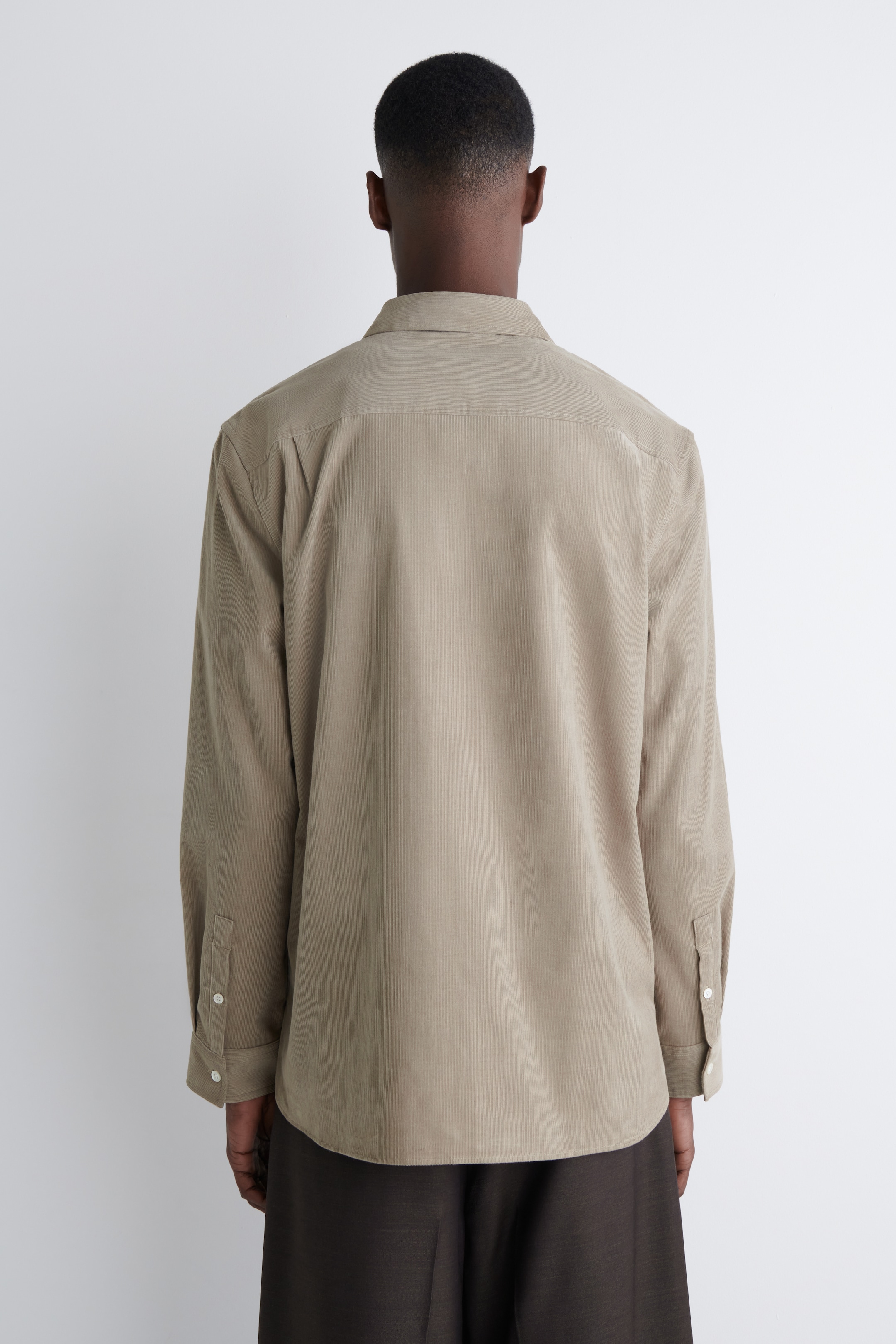 Agrandir l'image: Une chemise en velours côtelé beige est portée, vue de dos. Elle présente des manches longues avec des poignets boutonnés, un empiècement d'épaule et un ourlet arrondi, mettant en valeur sa coupe décontractée.