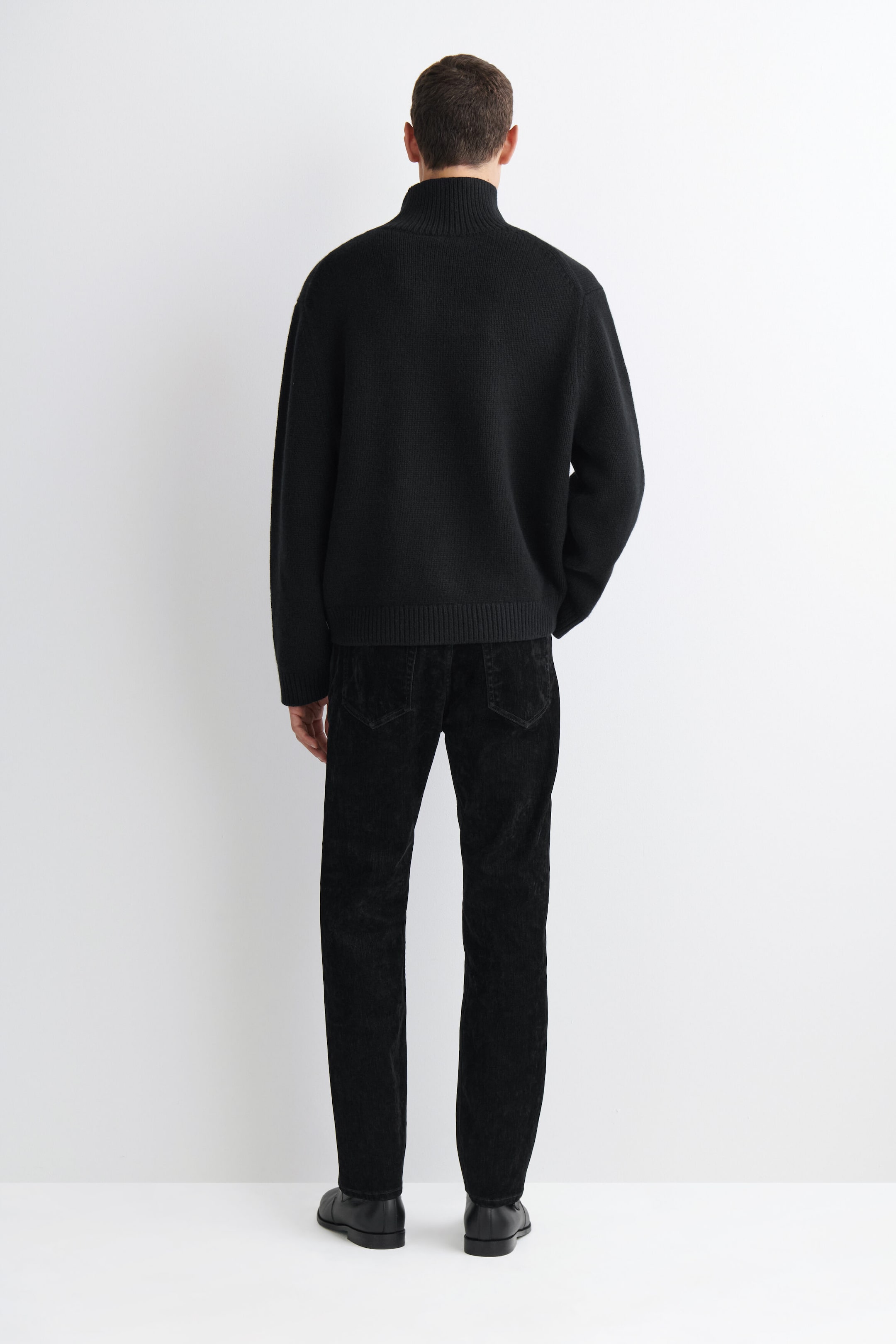 Los vaqueros Signature Flocked de corte recto se muestran por la parte trasera, destacando su ajuste recto y los dos bolsillos de parche traseros en un tejido flockado negro con textura. Se combinan con un jersey de punto negro de cuello alto y zapatos de piel negros.