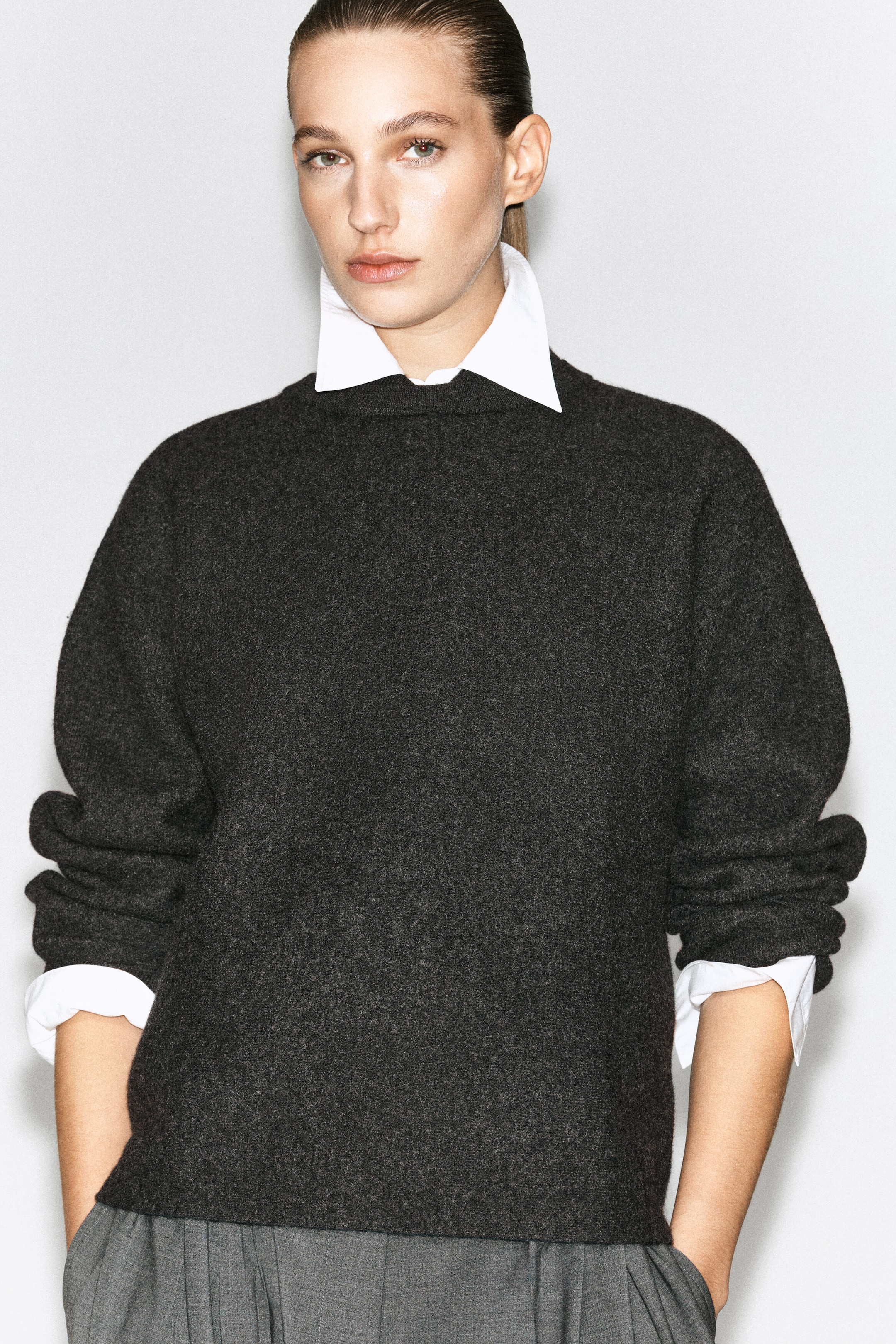 A dark gray boiled-wool sweater featuring a layered white pointed collar and matching white cuffs is worn by the model. The sweater is styled with gray pleated trousers.