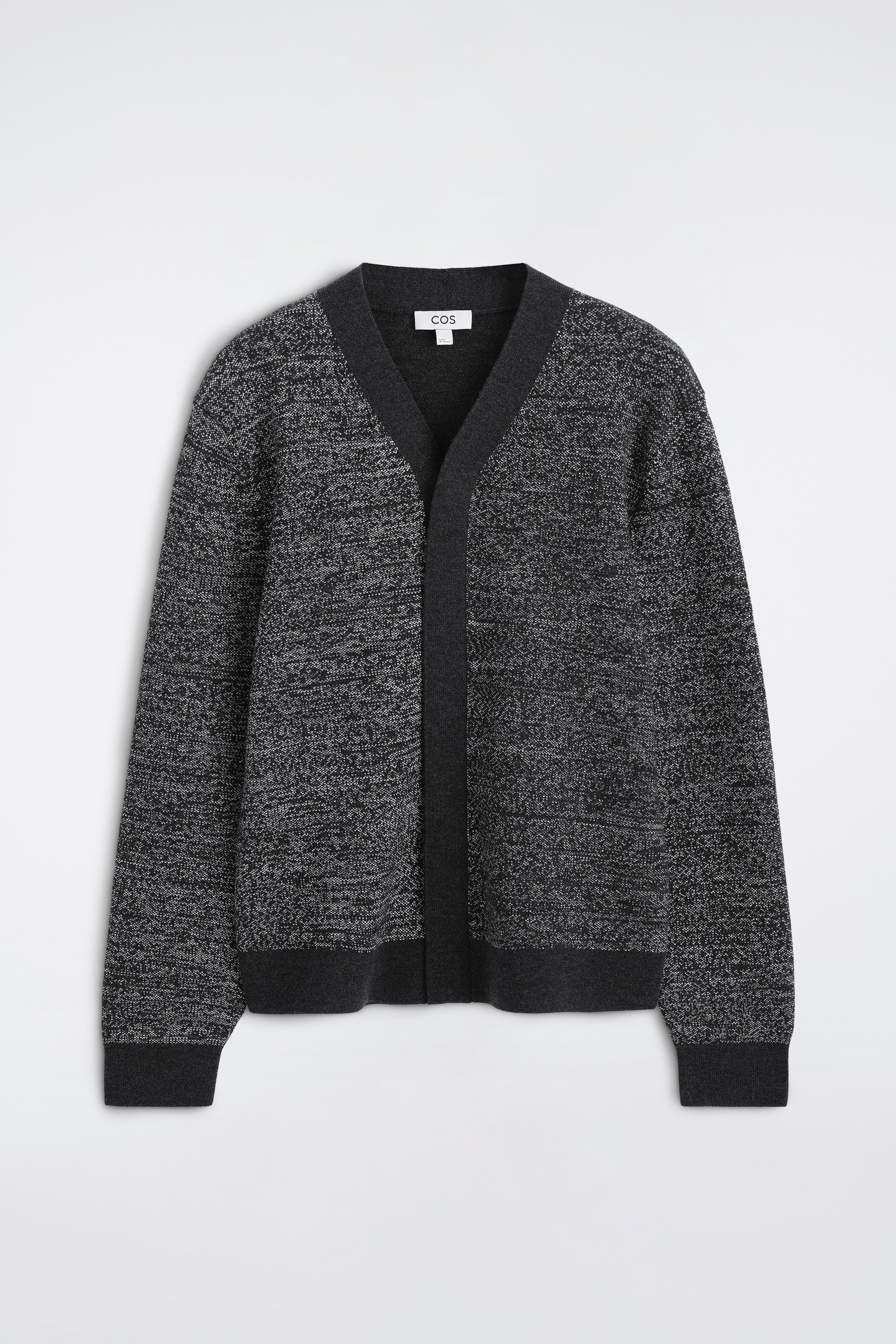 En gråmelerad stickad cardigan visas framifrån, med ett strukturerat grått- och vitt stickat mönster, en kontrasterande enfärgad mörkgrå V-ringad framkant, ribbade ärmslut och fåll. Cardiganen är lagerlagd över en vit T-shirt med rund hals och kombineras med svarta byxor för att framhäva dess lediga passform och design utan knappar.