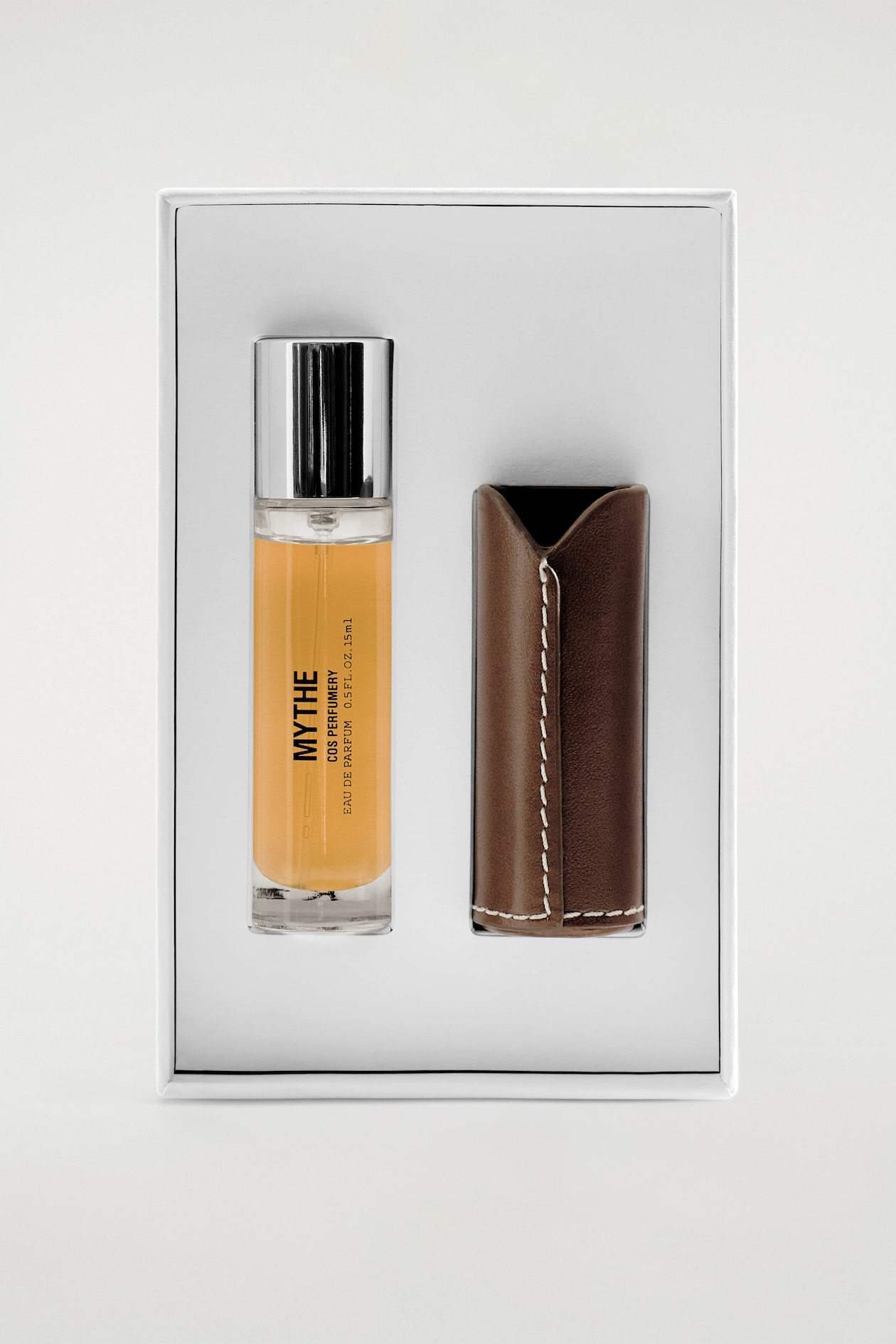 Shown from the front, a perfume set is resting inside a white presentation box, featuring a clear glass bottle filled with amber liquid and topped with a silver cap on the left, and a brown leather sleeve with white contrast stitching on the right.