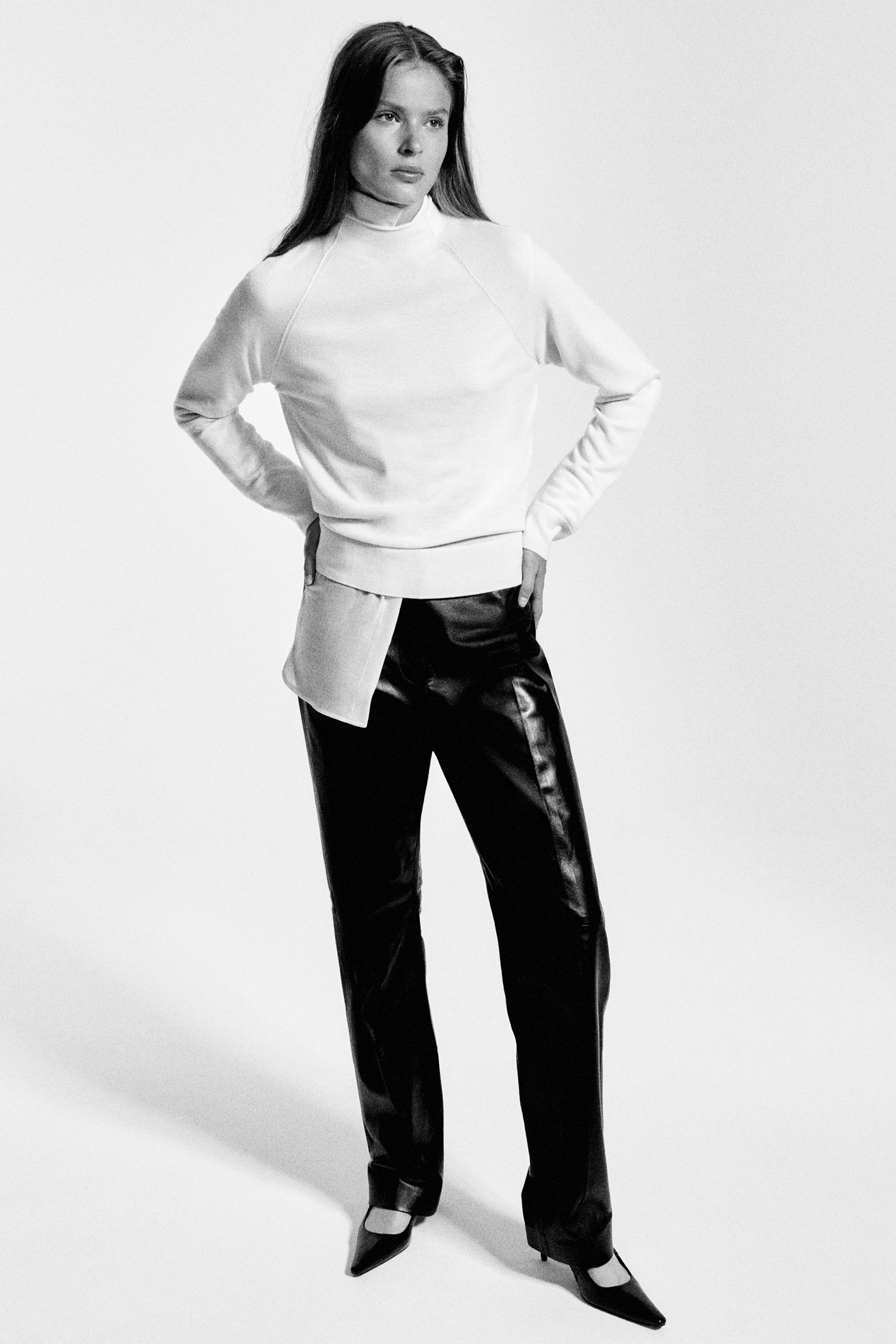 Grösseres Bild ansehen: Von vorne gezeigt verfügt der Merino-Woll-Pullover mit Funnel-Neck über lange Raglanärmel, einen hohen Kragen und einen gerippten Saum und ist über einem geschichteten Hemd mit Kragen gestylt. Dazu werden schwarze Leder-Hosen mit geradem Bein kombiniert, die einen dezenten Glanz sowie markante Bügelfalten entlang der Vorderseite der Beine aufweisen.