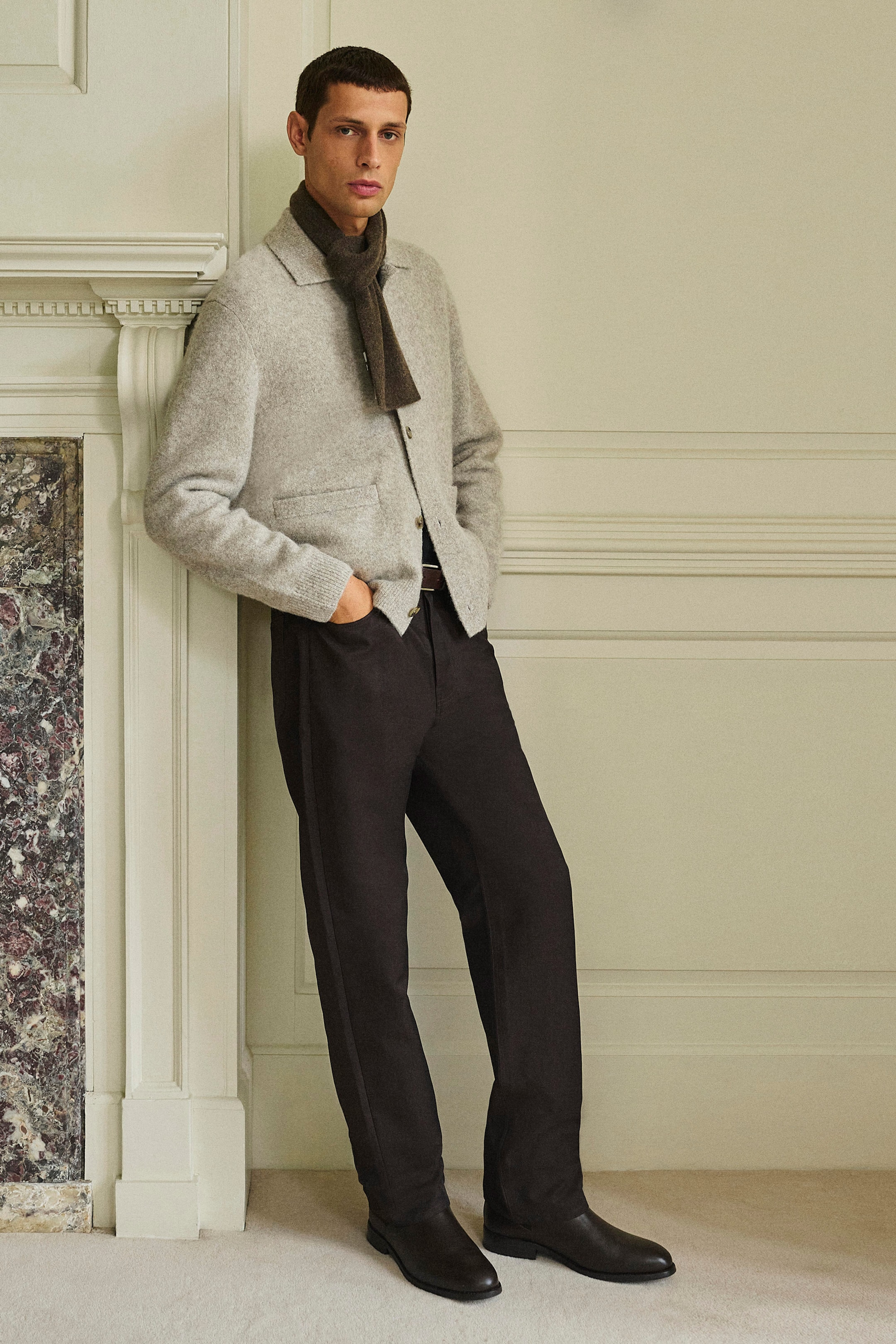A model is wearing a light grey textured collared cardigan featuring a classic collar, button-up front and front patch pockets, paired with a dark brown cashmere pull-through scarf looped around the neck. The look is completed with dark, straight-leg signature jeans, shown from the front to highlight their relaxed, fluid fit. A model is wearing a light grey textured collared cardigan featuring a classic collar, button-up front and front patch pockets, paired with a dark brown cashmere pull-through scarf looped around the neck. The look is completed with dark, straight-leg signature jeans, shown from the front to highlight their relaxed, fluid fit.
