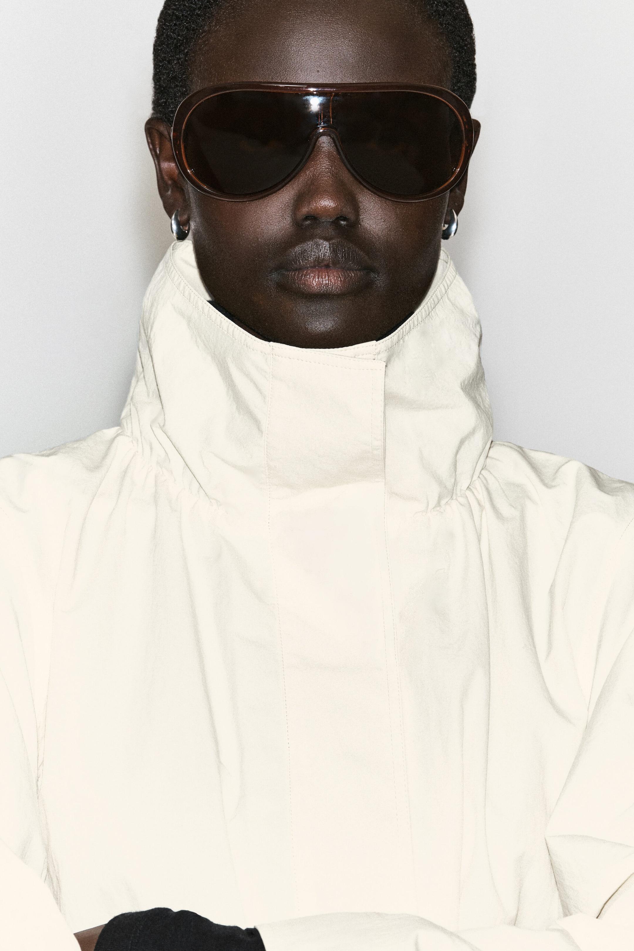 Une veste à col entonnoir en nylon blanc cassé est présentée en gros plan, de face, mettant en valeur son col montant structuré et haut qui couvre le cou. La veste présente une patte de boutonnage dissimulée à l’avant et de subtils fronces à la base du col. Une veste à col entonnoir en nylon blanc cassé est présentée en gros plan, de face, mettant en valeur son col montant structuré et haut qui couvre le cou. La veste présente une patte de boutonnage dissimulée à l’avant et de subtils fronces à la base du col.