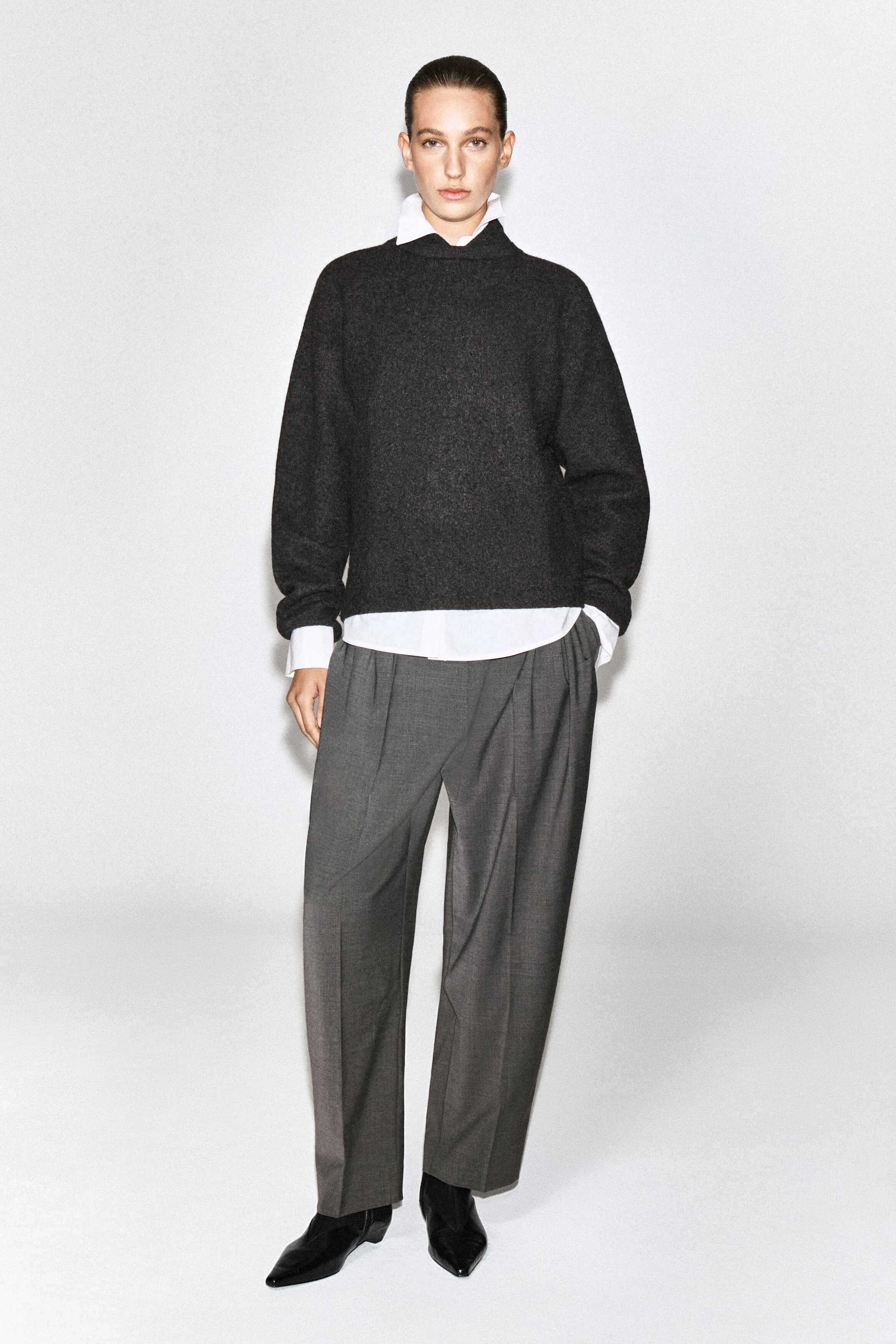 A dark gray boiled-wool sweater with layered white collar, cuffs, and hem is worn by the model. The sweater is styled with loose-fitting gray trousers and black pointed-toe shoes.