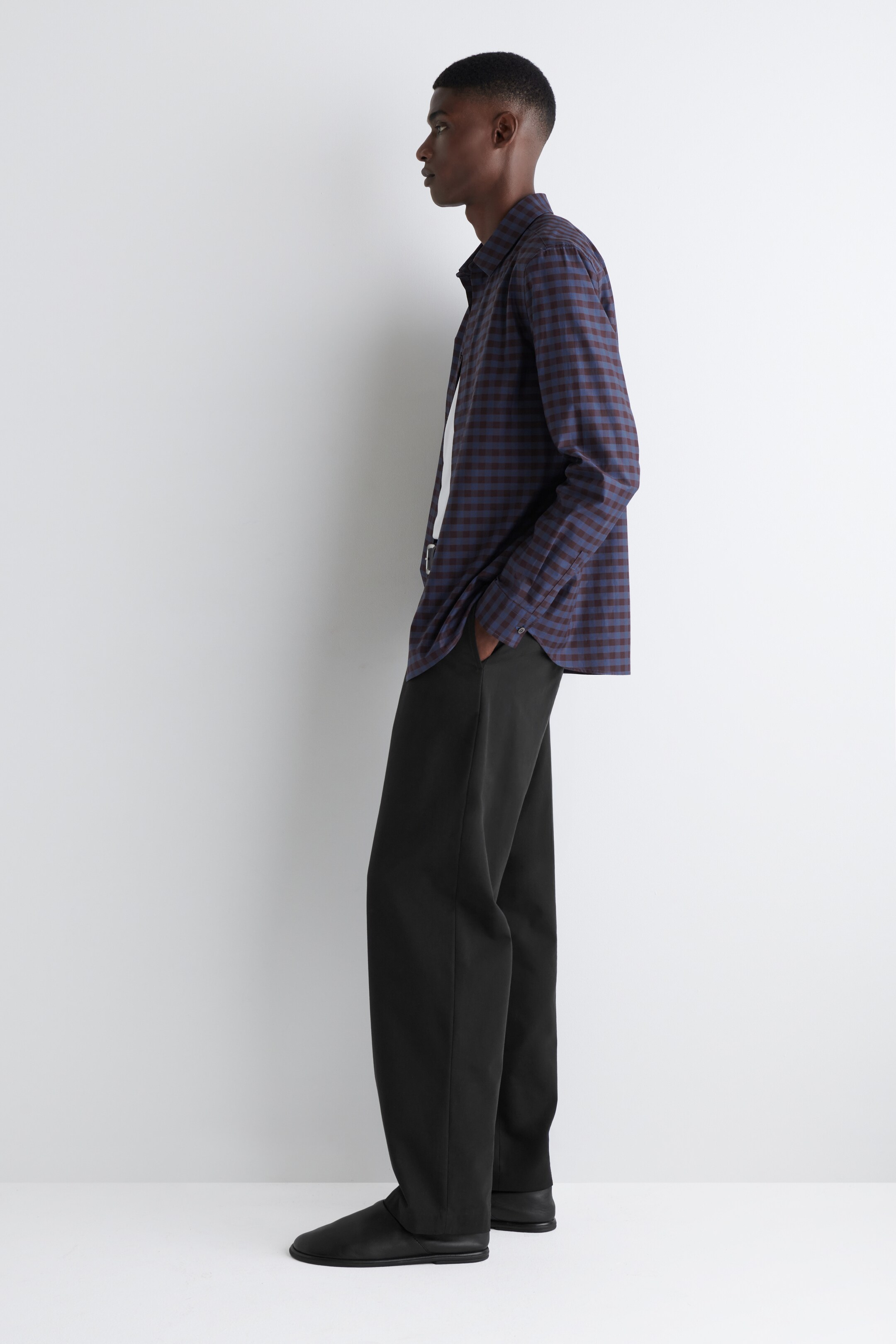 Ingrandisci l'immagine: Una camicia a maniche lunghe a quadretti gingham blu scuro e marrone rossiccio è indossata fuori dai pantaloni sopra una t-shirt bianca, abbinata a pantaloni neri a vestibilità rilassata e scarpe nere senza lacci, il tutto visto di lato.