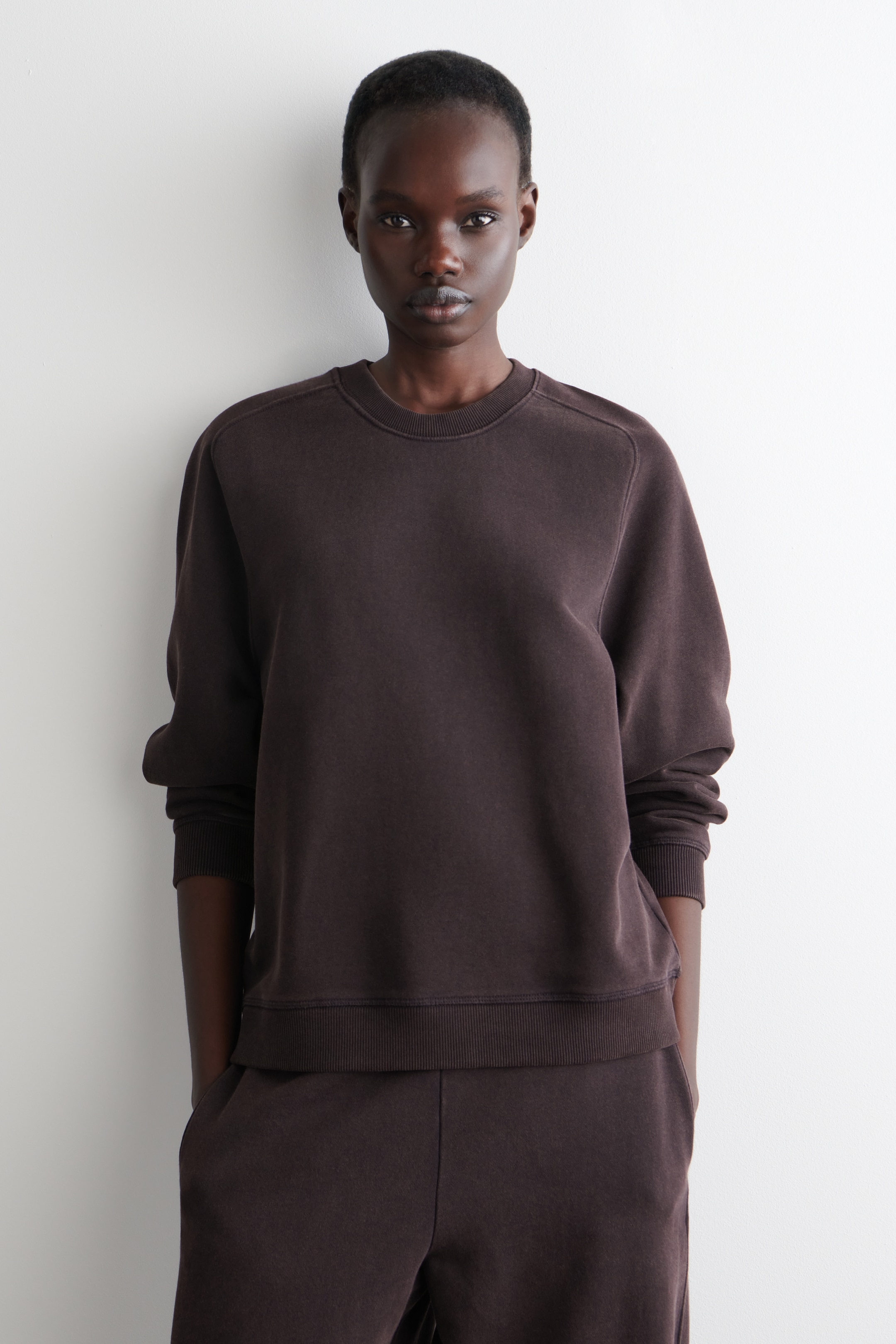 Agrandir l'image: Un sweat-shirt à col rond brun foncé est présenté de face, avec une coupe décontractée, des détails de couture visibles le long des épaules, et un col, des poignets et un ourlet côtelés. Il est assorti à un pantalon de survêtement brun foncé, complétant un ensemble de détente coordonné.