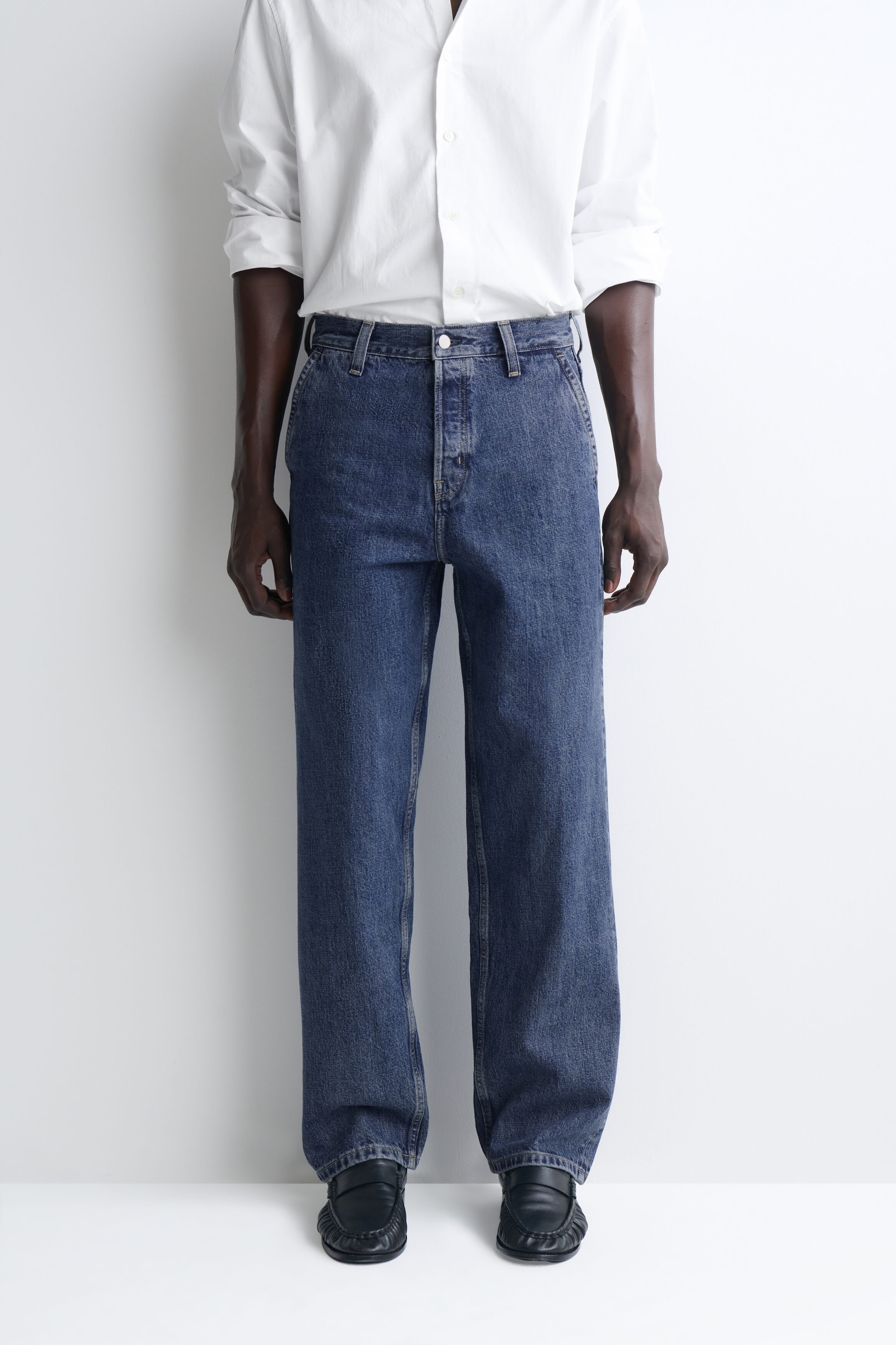 These relaxed-fit straight-leg denim trousers in a medium-blue wash are shown from the front, featuring a classic five-pocket design with belt loops and a single-button fastening. A tucked-in white shirt accentuates the waistline, while the straight legs fall loosely to pool slightly over black shoes. These relaxed-fit straight-leg denim trousers in a medium-blue wash are shown from the front, featuring a classic five-pocket design with belt loops and a single-button fastening. A tucked-in white shirt accentuates the waistline, while the straight legs fall loosely to pool slightly over black shoes.