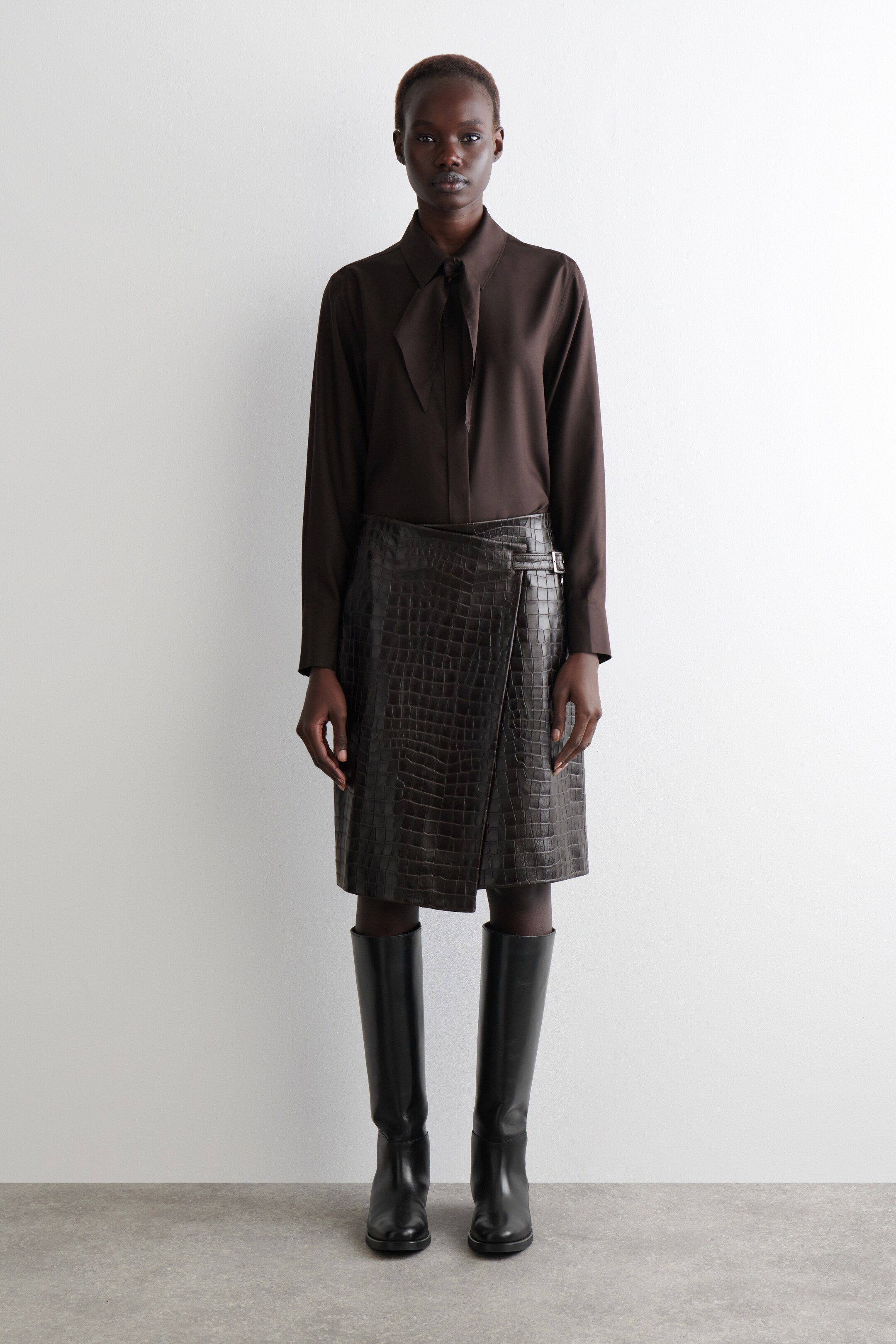 Agrandir l'image: Une blouse marron foncé à manches longues avec un col à nouer lâche est portée rentrée dans une jupe portefeuille marron foncé à motif crocodile, présentant une silhouette en A et un détail de boucle à la hanche. Des bottes en cuir noir montant jusqu'aux genoux complètent l'ensemble.