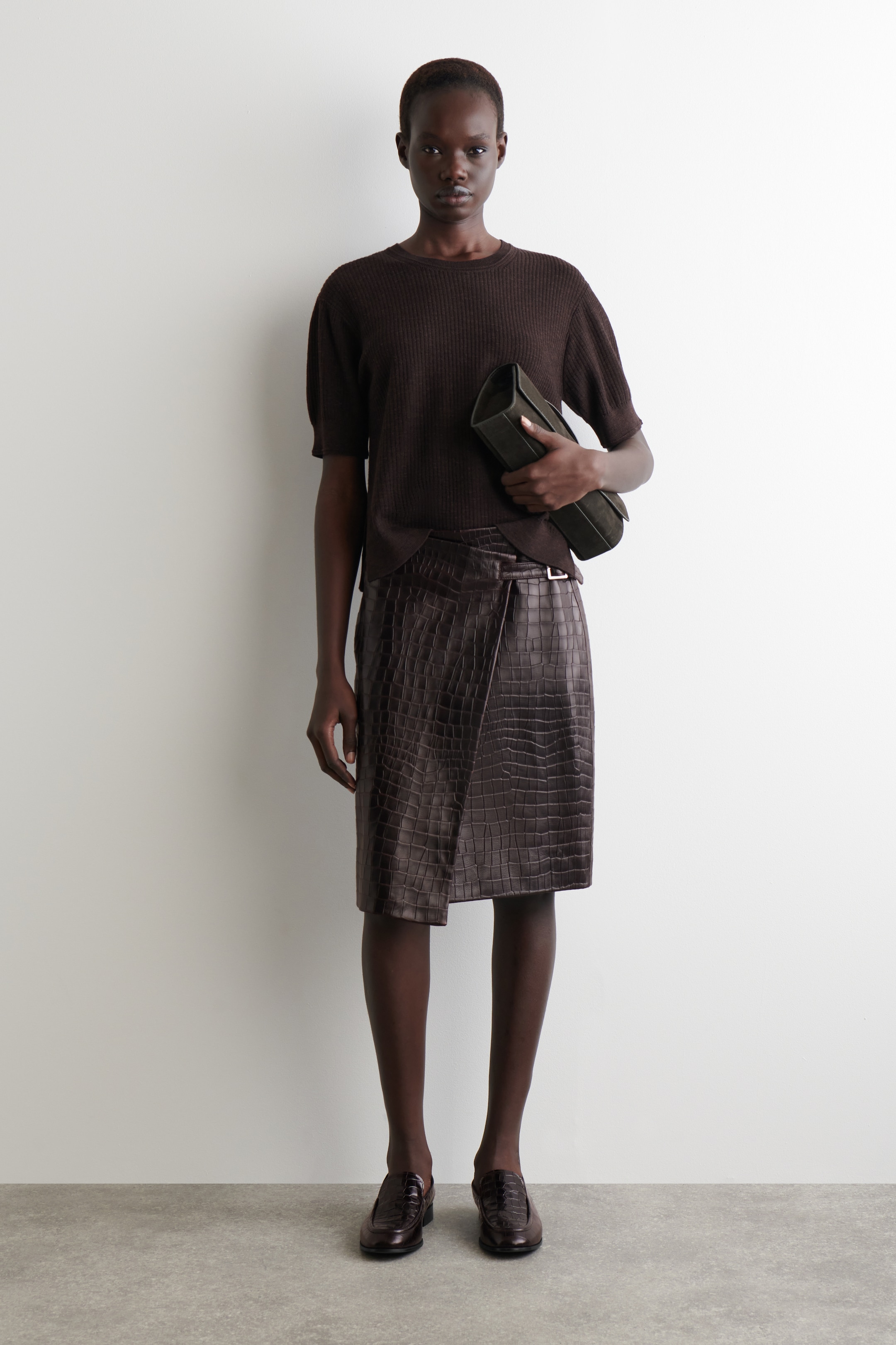 View larger image: A model is standing, wearing a dark brown, short-sleeved, ribbed knit top with a crew neck and a dark brown, crocodile-embossed wrap skirt featuring a side buckle detail, reaching just below the knee. She is holding a dark olive clutch bag under her arm and is wearing dark brown, crocodile-embossed loafers.