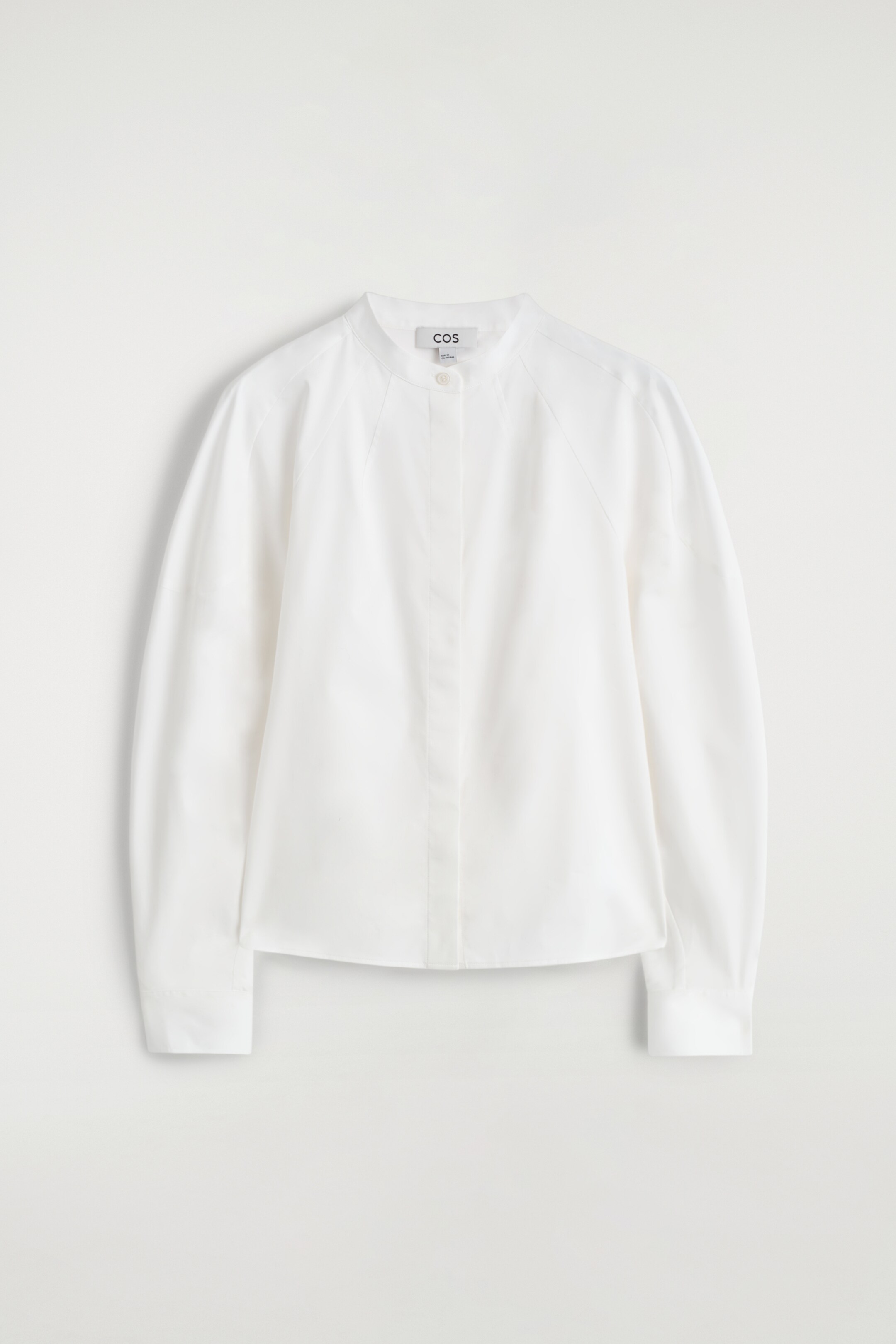 View larger image: A white shirt is displayed FRONT, featuring a collarless neckline, raglan shoulders, and a hidden button placket with a single visible top button, while long sleeves are finishing with cuffs.