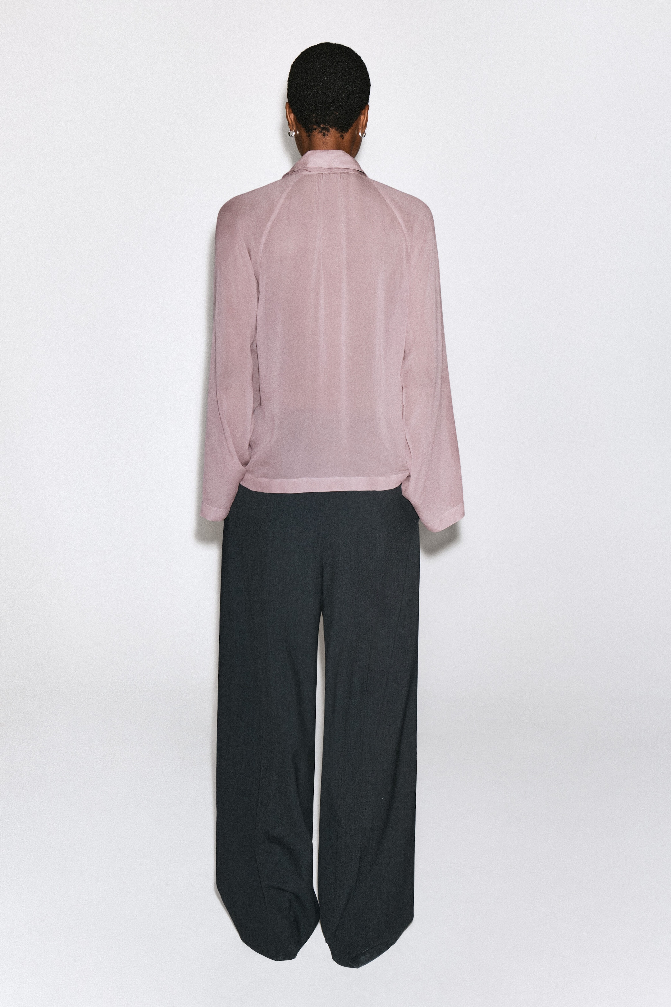 Le haut fluide avec détail foulard, en rose poudré, est porté par le mannequin et met en valeur son tombé décontracté, son col montant et ses manches longues vus de dos. Le haut est associé à un pantalon large gris foncé.