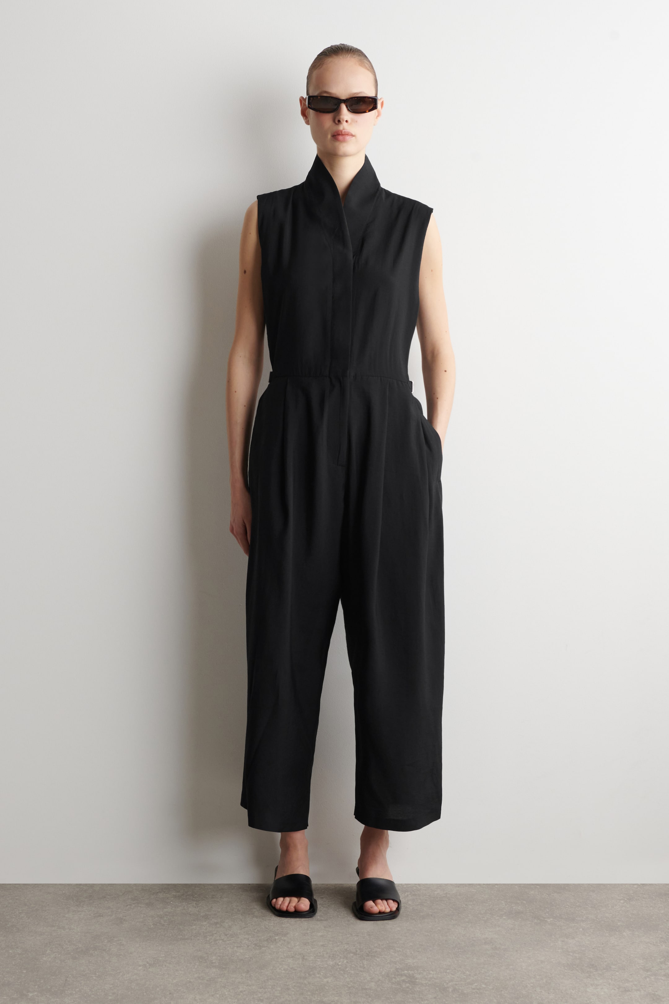 View larger image: A black sleeveless jumpsuit featuring a stand collar and a front placket. It is designed with a cinched waist and continues into wide-leg, cropped trousers with visible side pockets.