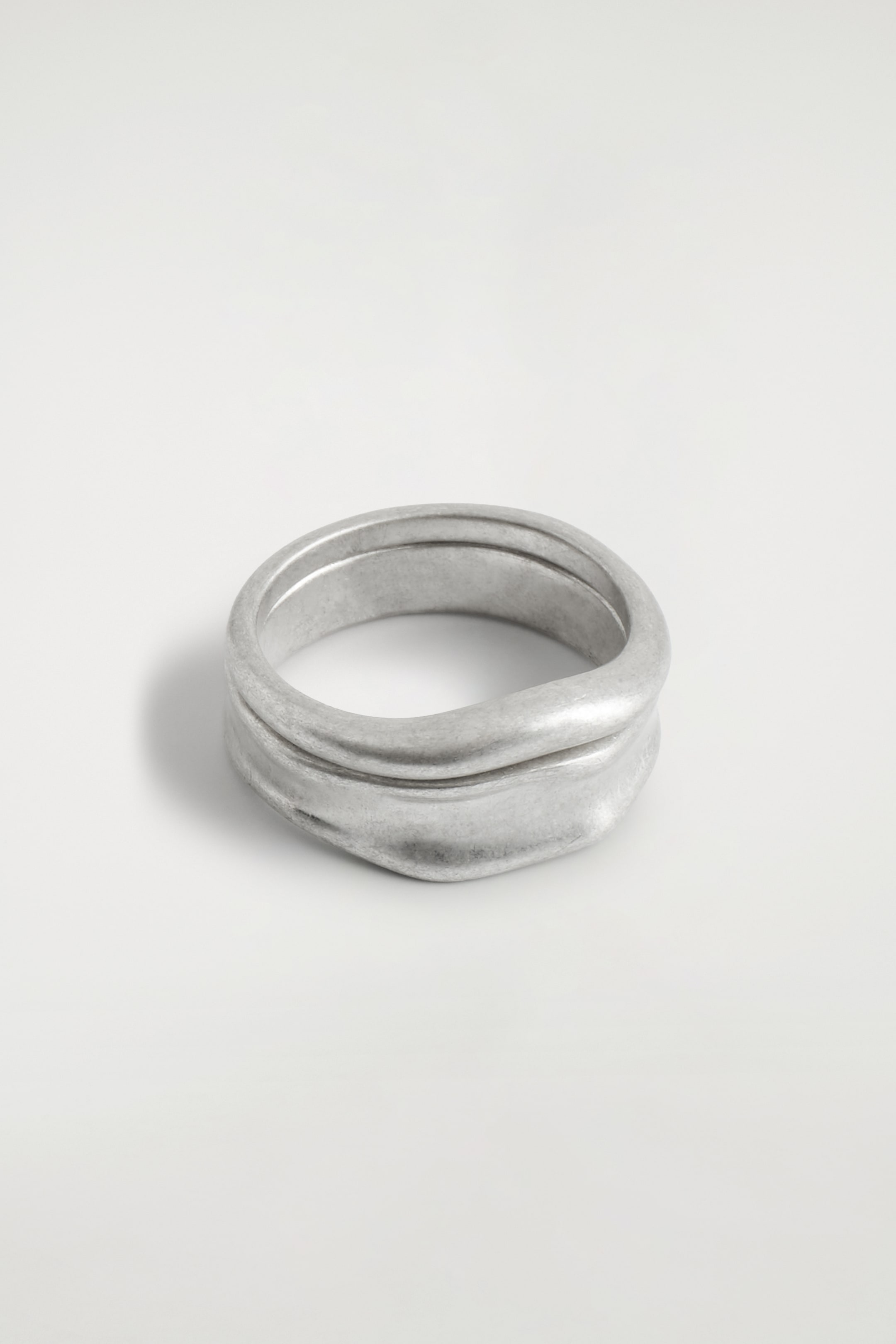 View larger image: A silver-toned ring is presenting from a slightly elevated angle, its wide band featuring an irregular, molten silhouette with a subtly tarnished, matte finish, and appearing to fold over itself in a wavy pattern.