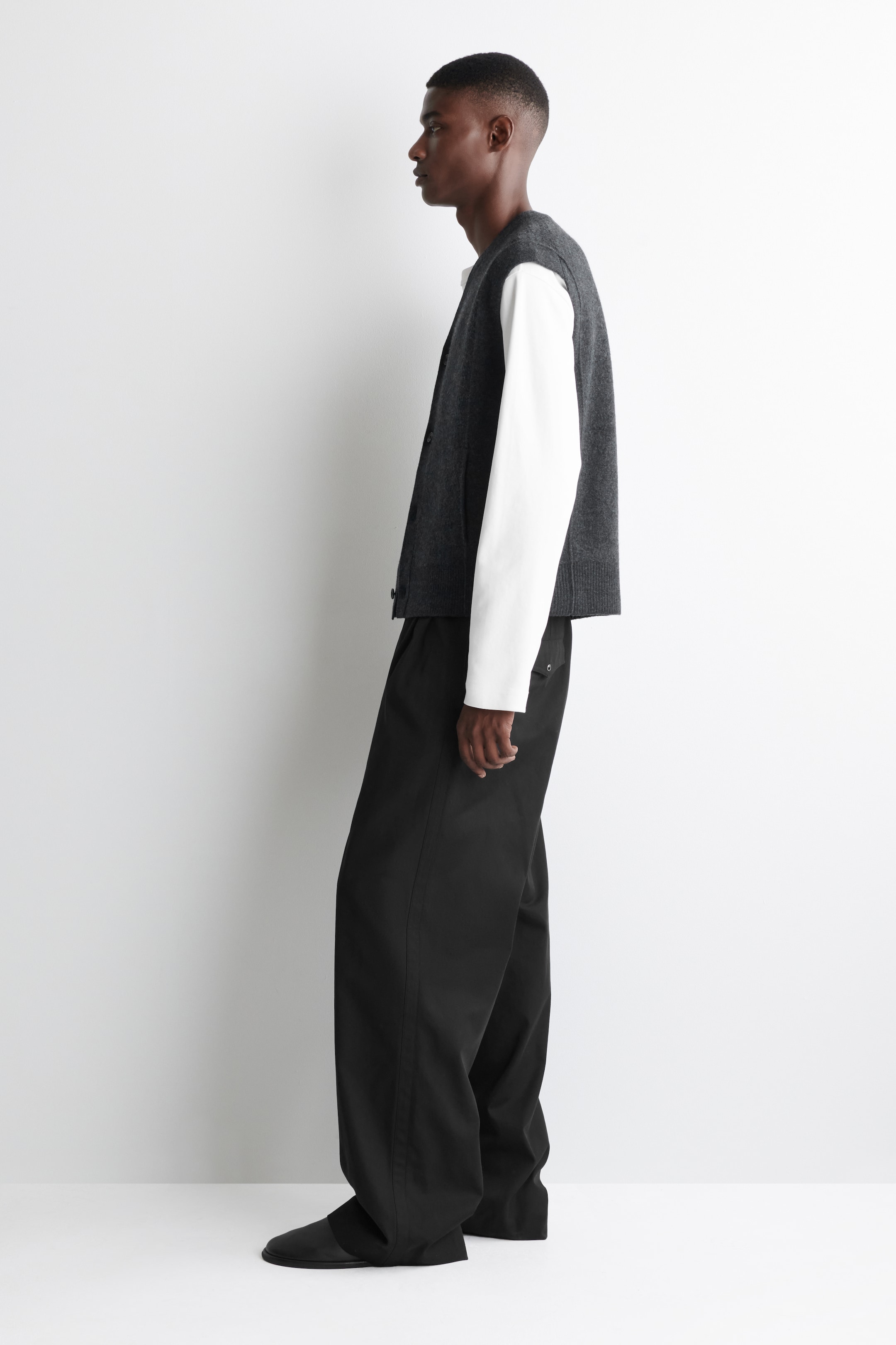 View larger image: A dark grey knit V-neck sweater vest, featuring a button-front closure, is being worn over a white long-sleeved shirt. This outfit is presented from a side profile, with black wide-leg trousers pooling slightly over black flat shoes.