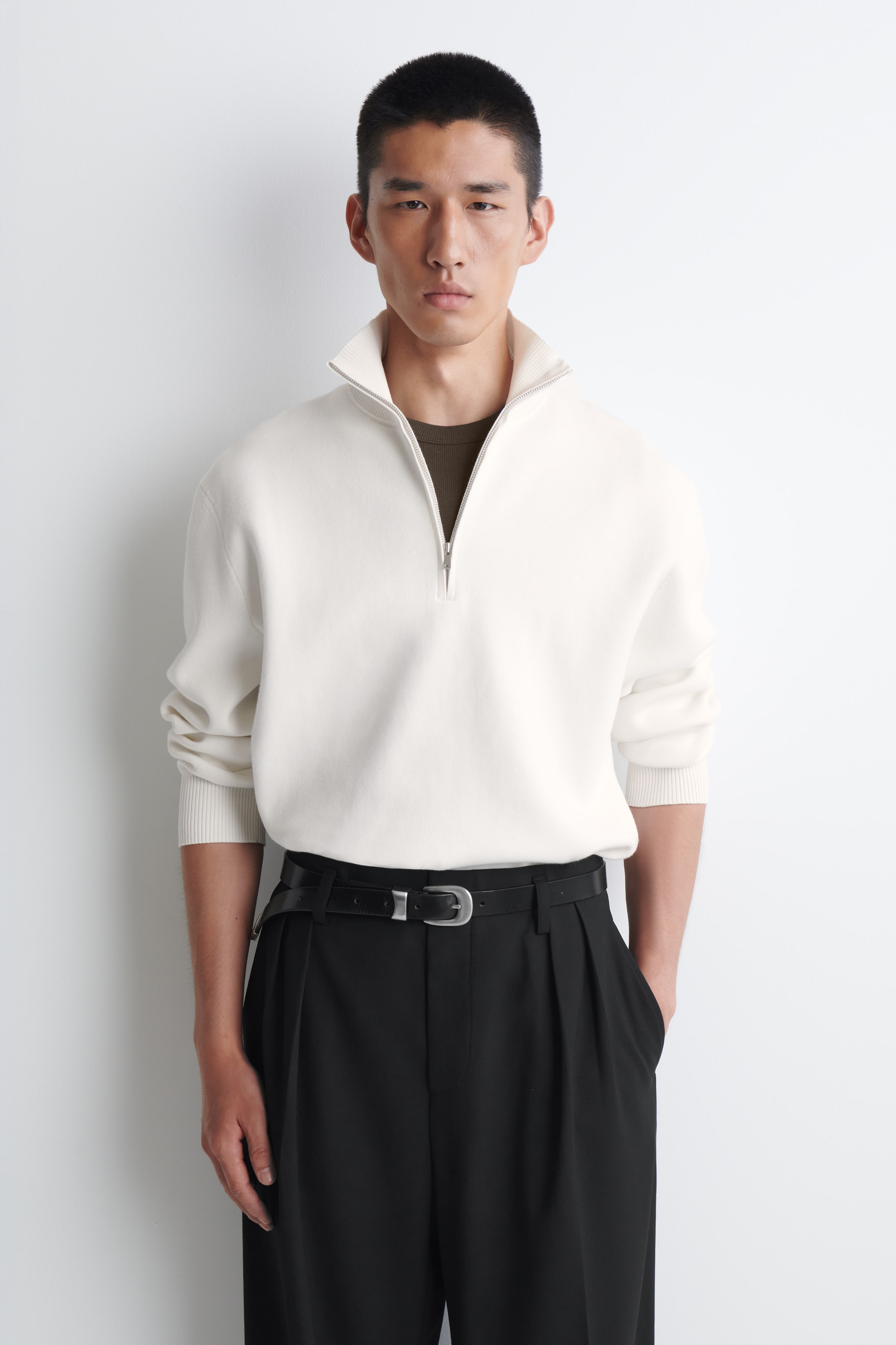 Agrandir l'image: Un pull couleur crème présente un col cheminée côtelé avec une fermeture à demi-zip effet argenté sur le devant. Il affiche une coupe classique avec des manches longues et des poignets côtelés, et est porté par-dessus un t-shirt foncé.