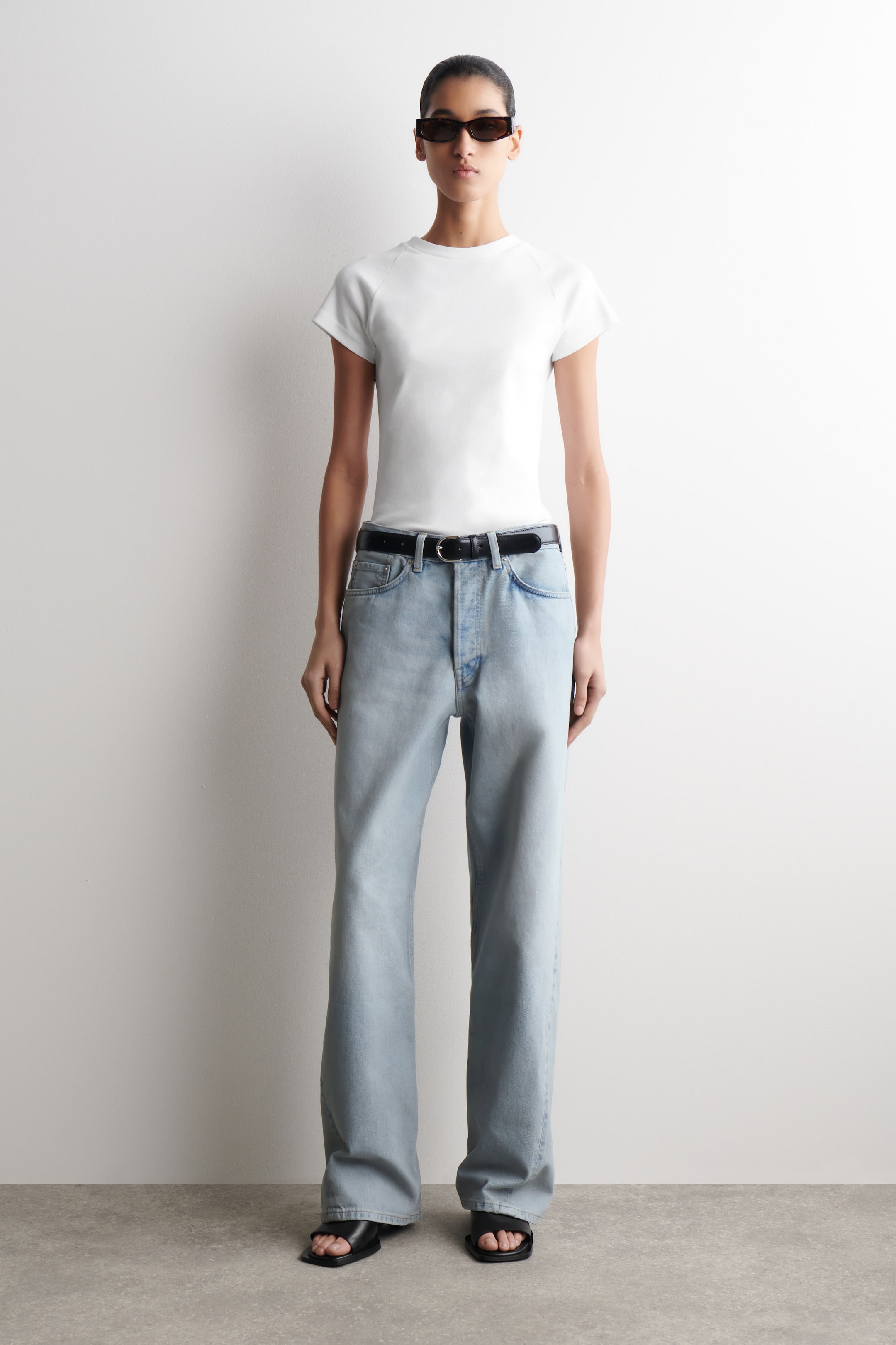 Agrandir l'image: Jean à large jambe et délavage clair, présenté de face, avec une taille haute, une fermeture à boutons et des poches avant, stylisé avec une ceinture noire et un t-shirt blanc rentré.