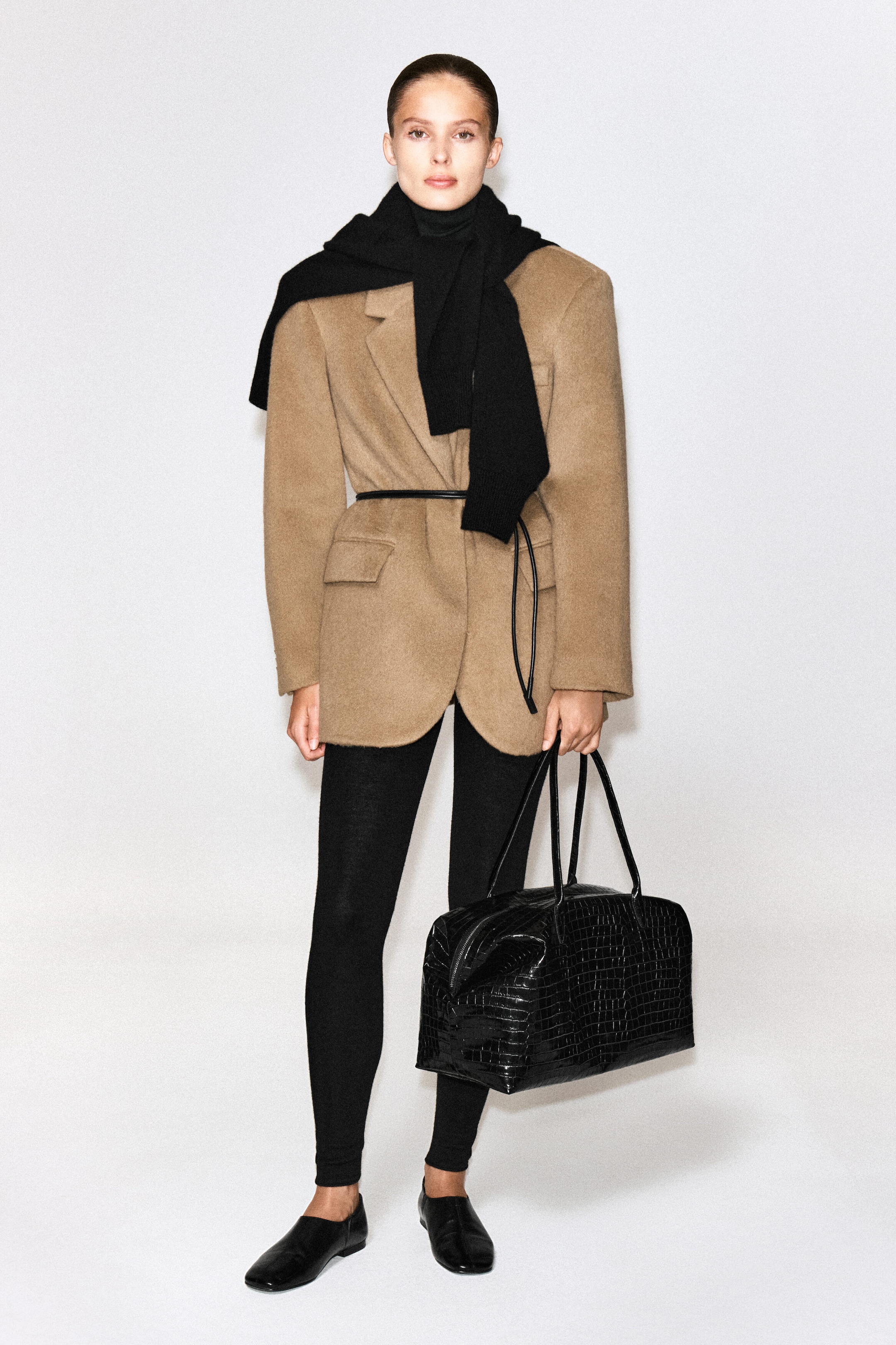 The Frieze Bowling Bag in black croc-effect leather is being carried by the model, who is holding it by its two long top handles. The bag features a structured, rectangular silhouette with a glossy, crocodile-embossed texture. The Frieze Bowling Bag in black croc-effect leather is being carried by the model, who is holding it by its two long top handles. The bag features a structured, rectangular silhouette with a glossy, crocodile-embossed texture.
