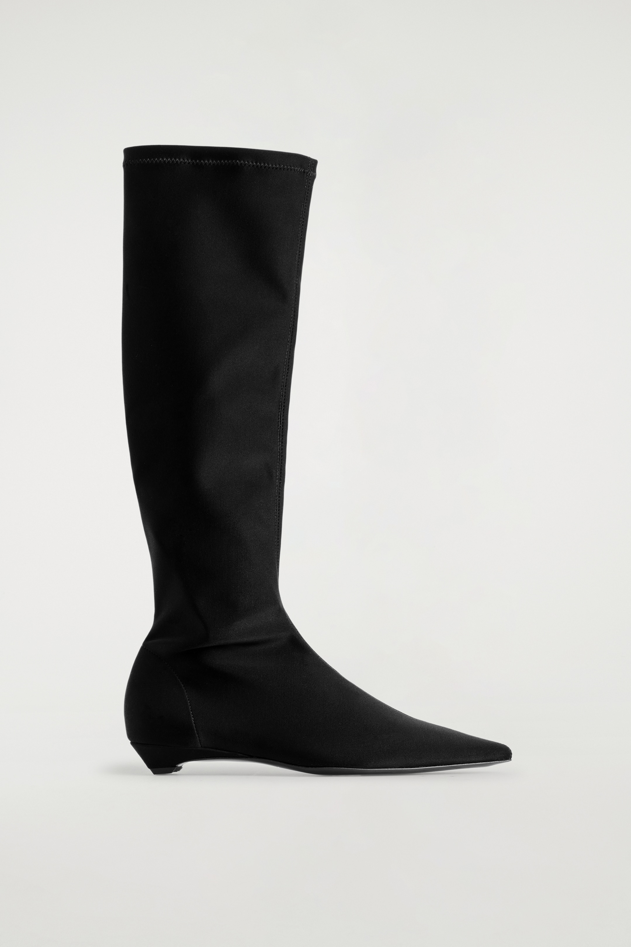 View larger image: The black pointed scuba knee sock boot is standing, facing right, featuring a sleek, form-fitting knee-high shaft, a sharply pointed toe, and a low heel.