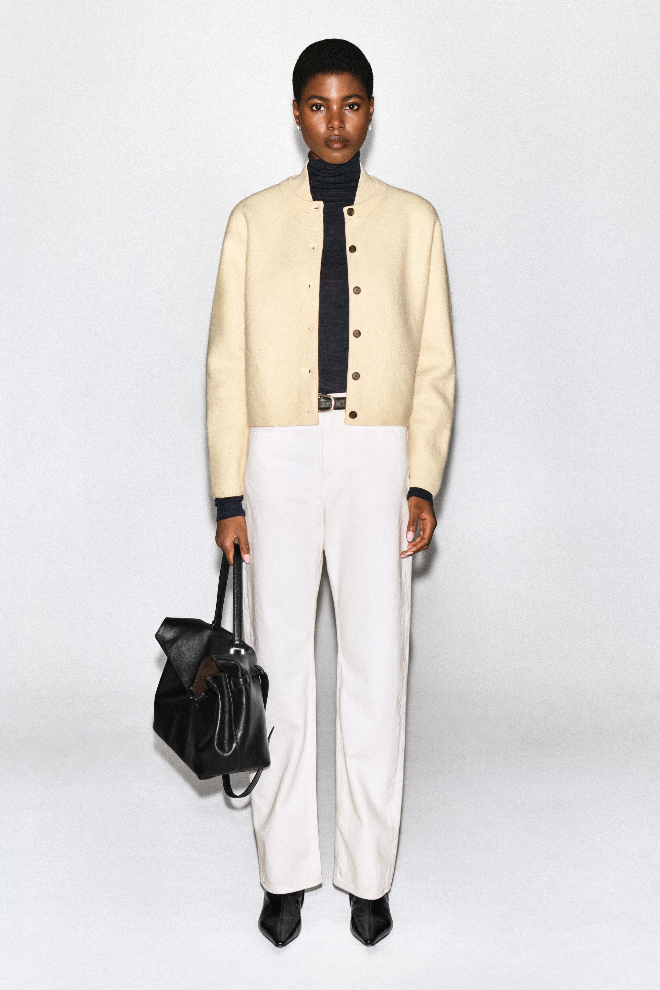 Un cardigan en laine bouillie de couleur crème, doté d’un col de style bomber et d’une fermeture boutonnée sur le devant, est porté ouvert par le mannequin. Le cardigan est superposé à un col roulé gris foncé et agencé avec un pantalon blanc ainsi qu’un sac à main en cuir noir tenu par le mannequin. Un cardigan en laine bouillie de couleur crème, doté d’un col de style bomber et d’une fermeture boutonnée sur le devant, est porté ouvert par le mannequin. Le cardigan est superposé à un col roulé gris foncé et agencé avec un pantalon blanc ainsi qu’un sac à main en cuir noir tenu par le mannequin.