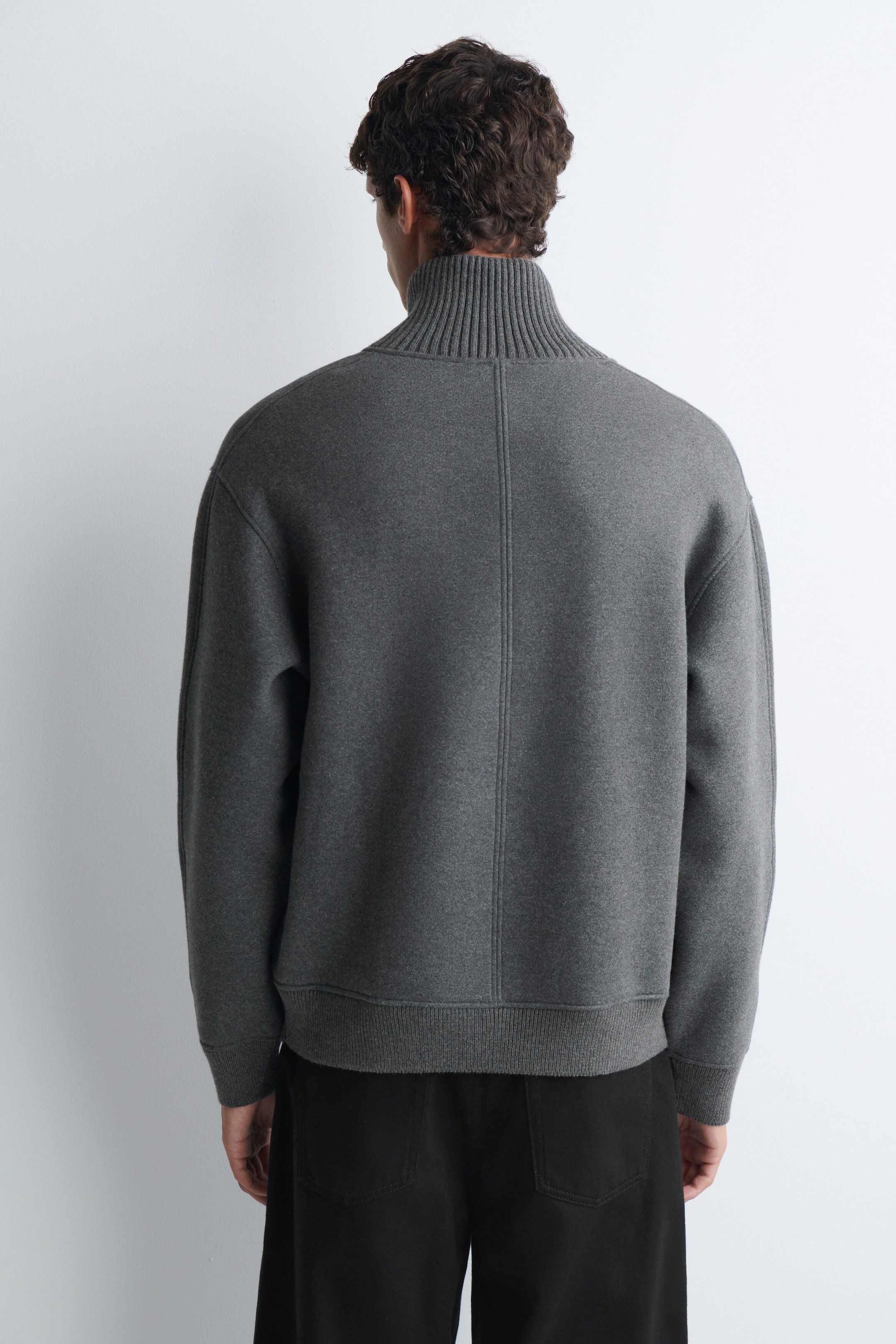 A cotton-wool jersey zip-up jumper in grey is being worn by the model, showcasing the back design with a high ribbed collar, a central vertical seam, and ribbed cuffs and hem. The jumper is styled with black trousers. A cotton-wool jersey zip-up jumper in grey is being worn by the model, showcasing the back design with a high ribbed collar, a central vertical seam, and ribbed cuffs and hem. The jumper is styled with black trousers.
