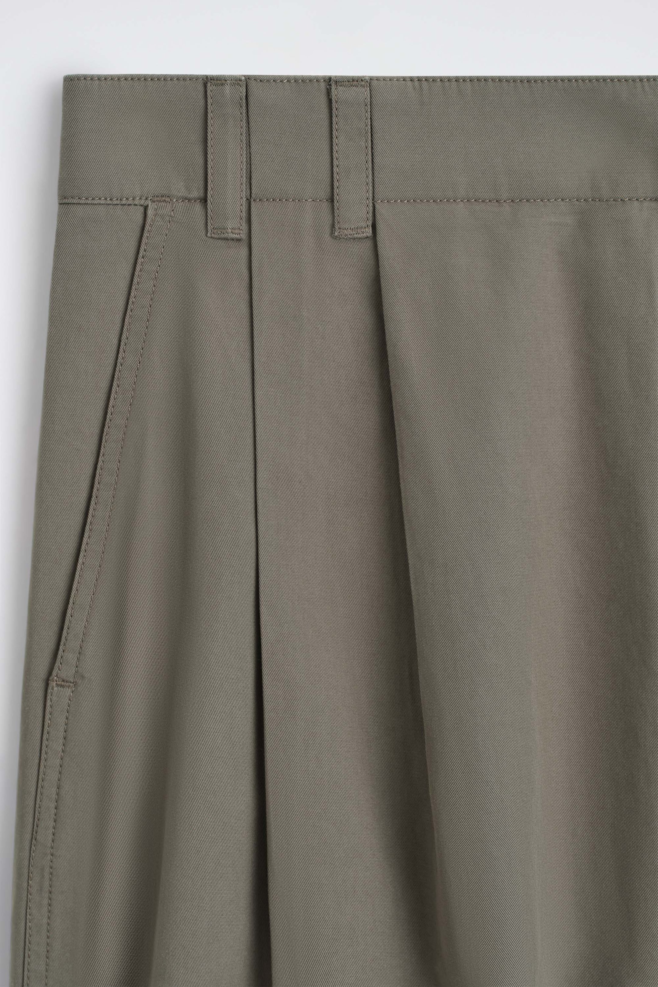 Die maßgeschneiderten, plissierten Baumwollshorts sind in einer nahen Frontalansicht des olivgrauen Twill-Stoffs zu sehen und präsentieren einen strukturierten Bund mit Gürtelschlaufen. Eine tiefe vertikale Falte und eine seitliche Schrägtasche mit detaillierten Steppnähten prägen die vordere Konstruktion.