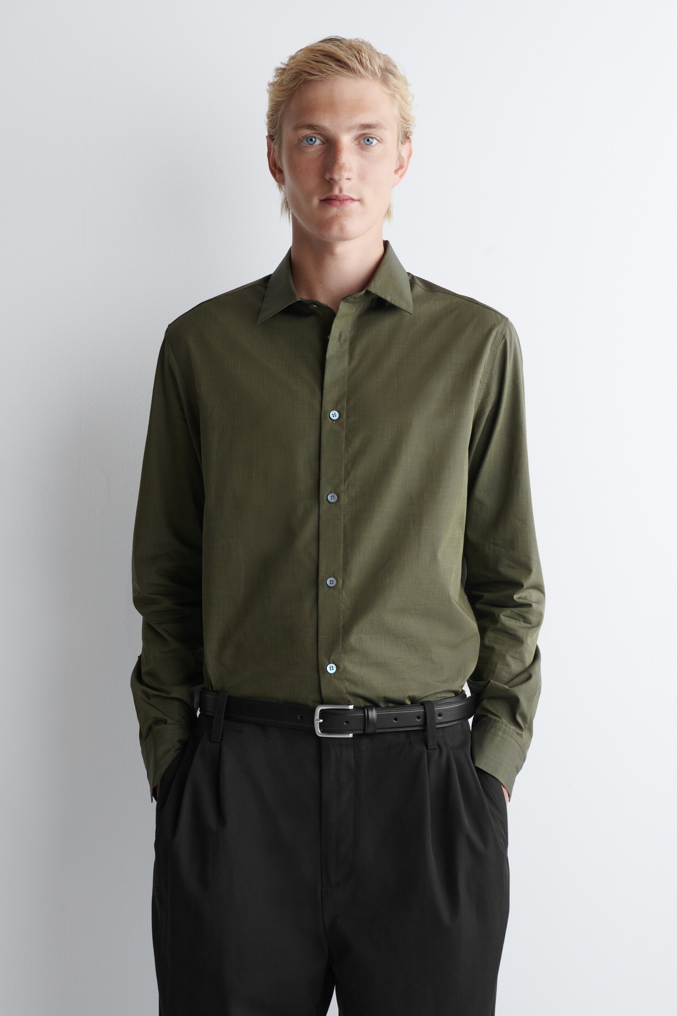 Ver imagen más grande: Una camisa verde oliva de manga larga se presenta de frente. Presenta una textura tejida sutil, un cuello en punta, una tapeta de botones completa con botones gris claro y puños con botones. La camisa está metida dentro de unos pantalones negros.