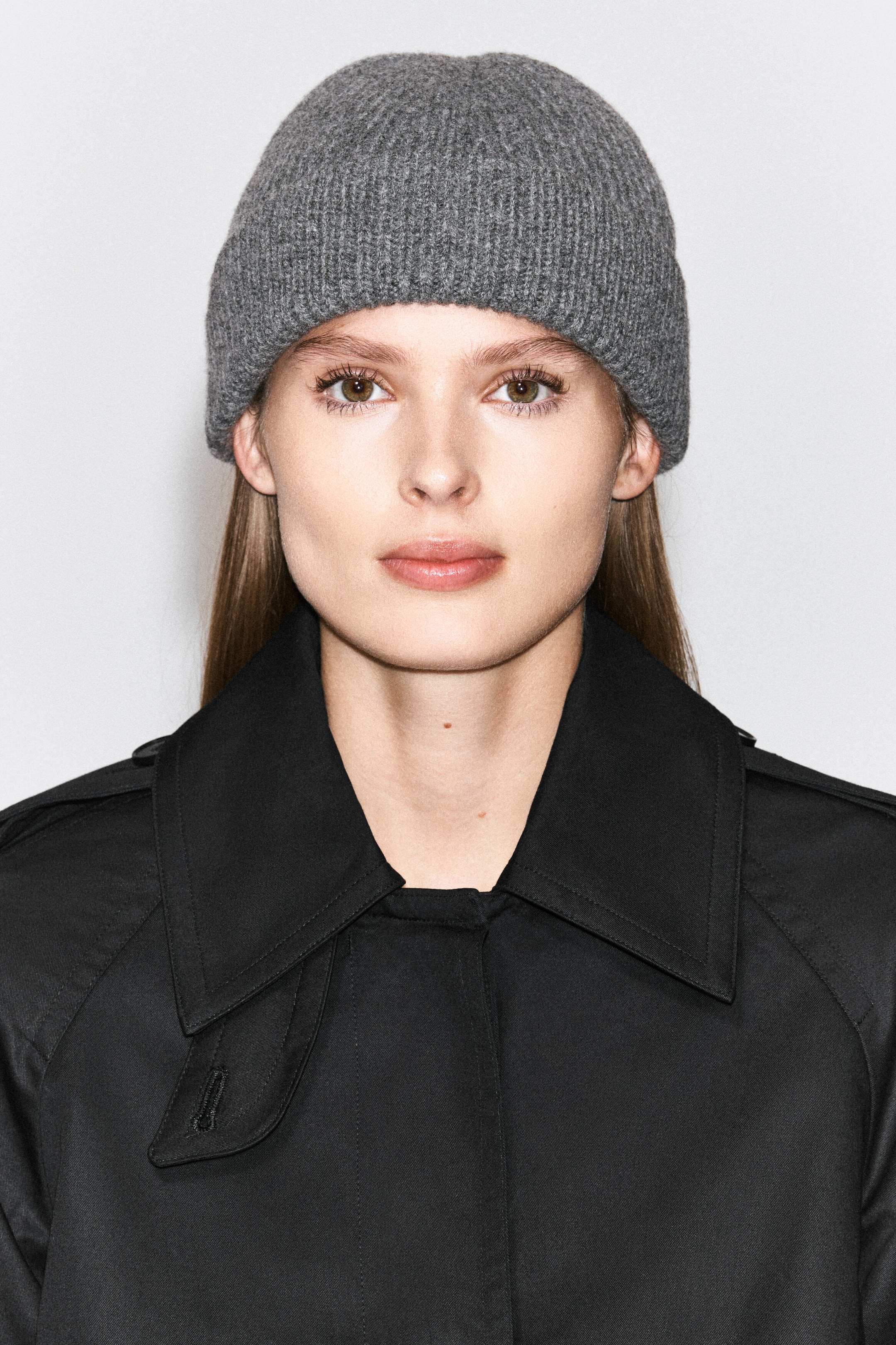 A gray ribbed knit beanie is being worn by the model, covering the upper forehead. The beanie is styled with a black trench coat featuring a wide, structured collar. A gray ribbed knit beanie is being worn by the model, covering the upper forehead. The beanie is styled with a black trench coat featuring a wide, structured collar.