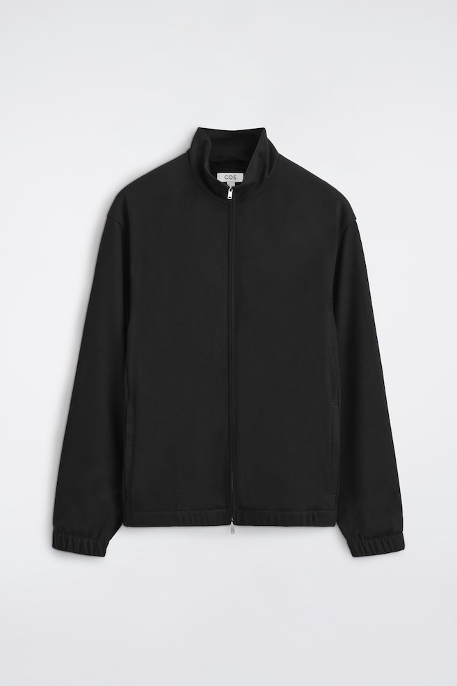 Diese schwarze Blousonjacke aus Jersey mit Stehkragen und ReiÃverschluss ist von vorne zu sehen und zeigt einen hohen Stehkragen, einen durchgehenden silberfarbenen ReiÃverschluss sowie dezente paspelierte Seitentaschen. Die langen Ãrmel und der Saum sind mit gerafften, elastischen Details versehen, die eine lässige Blouson-Silhouette erzeugen. - 1