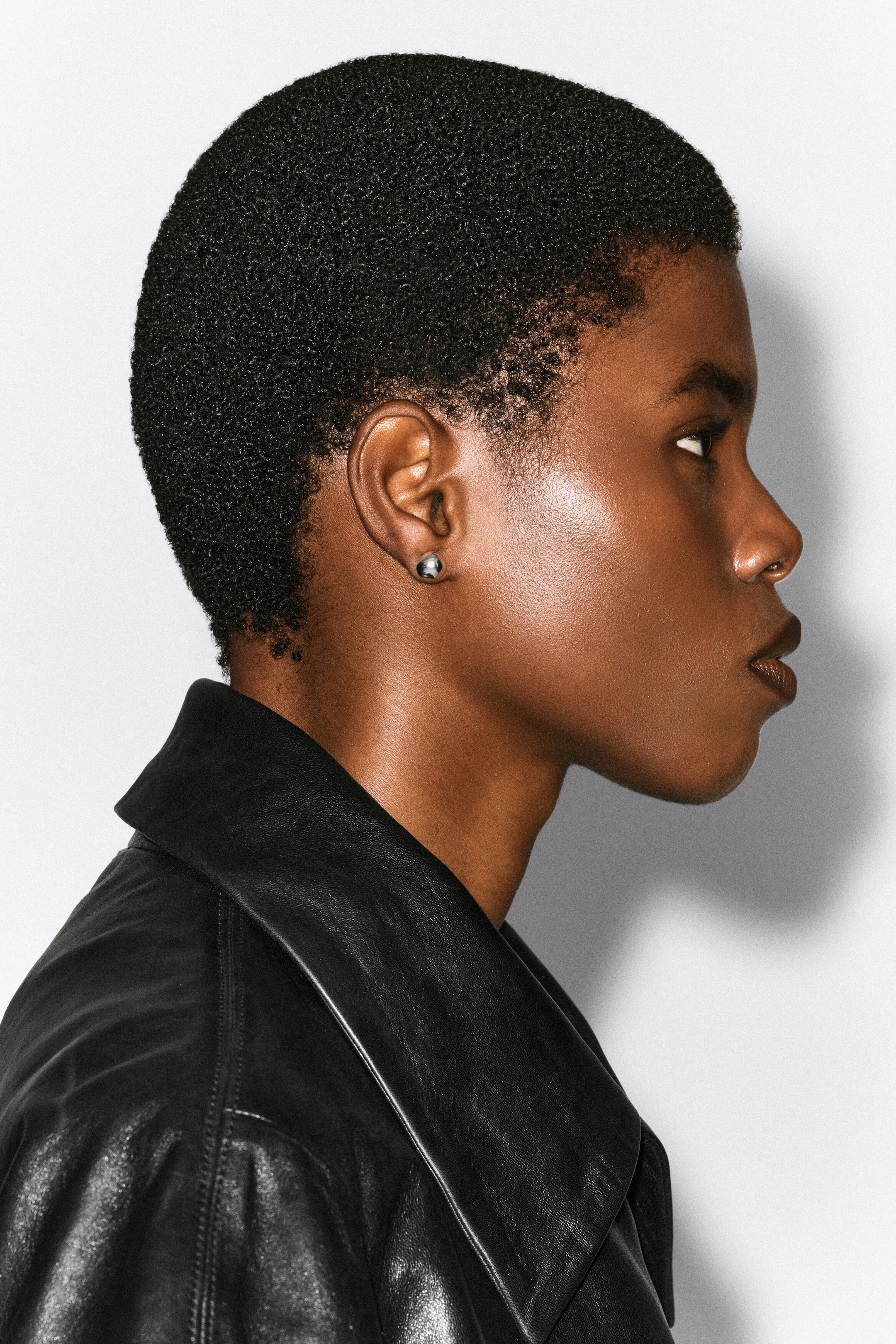 A small, spherical silver-tone earring is being worn on the model's earlobe, shown in profile and wearing a black leather jacket.