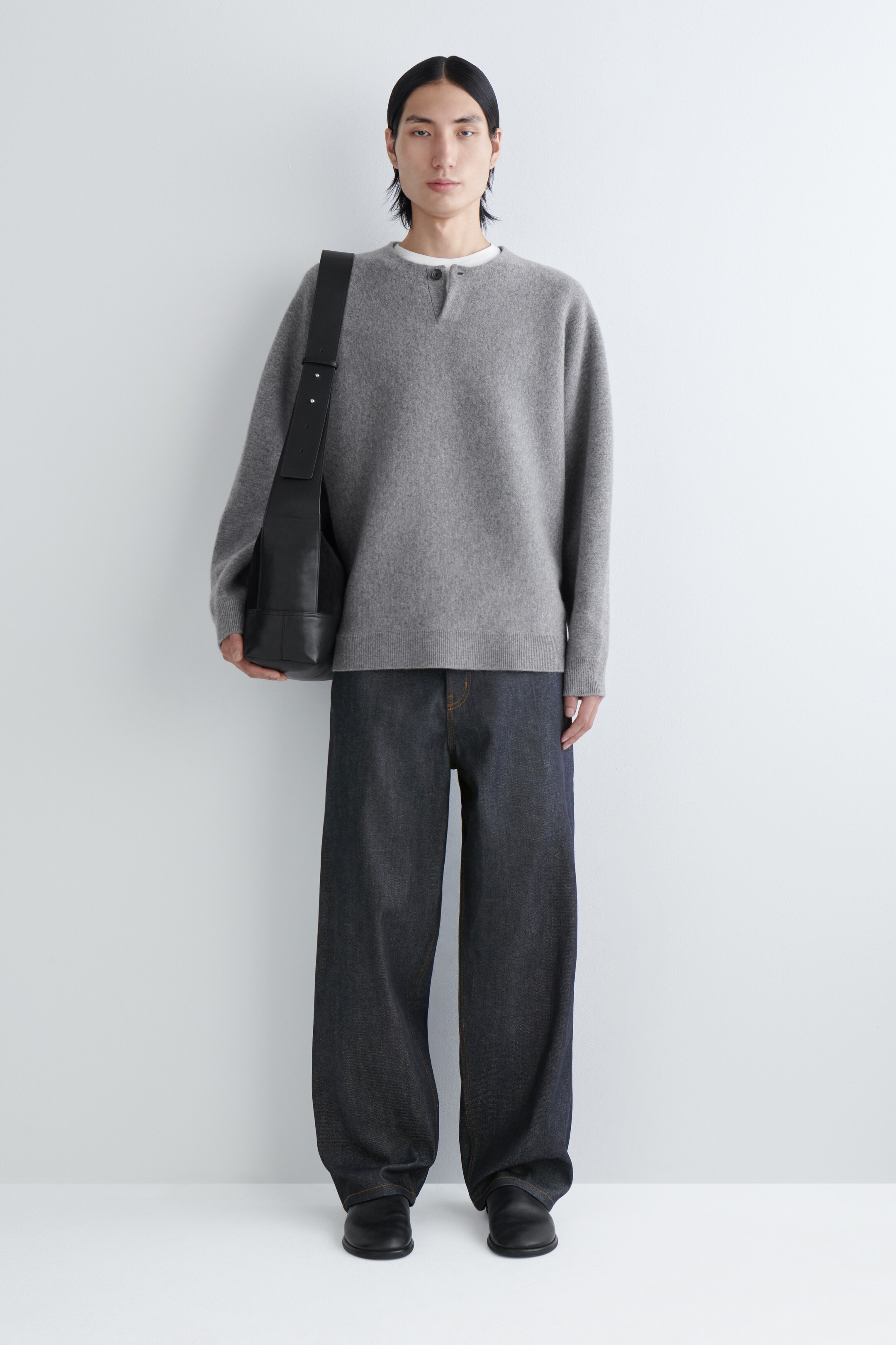 Agrandir l'image: Un pull henley à manches longues gris clair est porté par-dessus un t-shirt à col rond blanc, présenté de face. Un jean large indigo foncé avec des surpiqûres orange contrastantes est porté, apparaissant en pleine longueur et s'accumulant légèrement aux chevilles. Un sac à bandoulière en cuir noir avec une large sangle plate est suspendu à l'épaule gauche, et des chaussures noires à enfiler sont visibles.