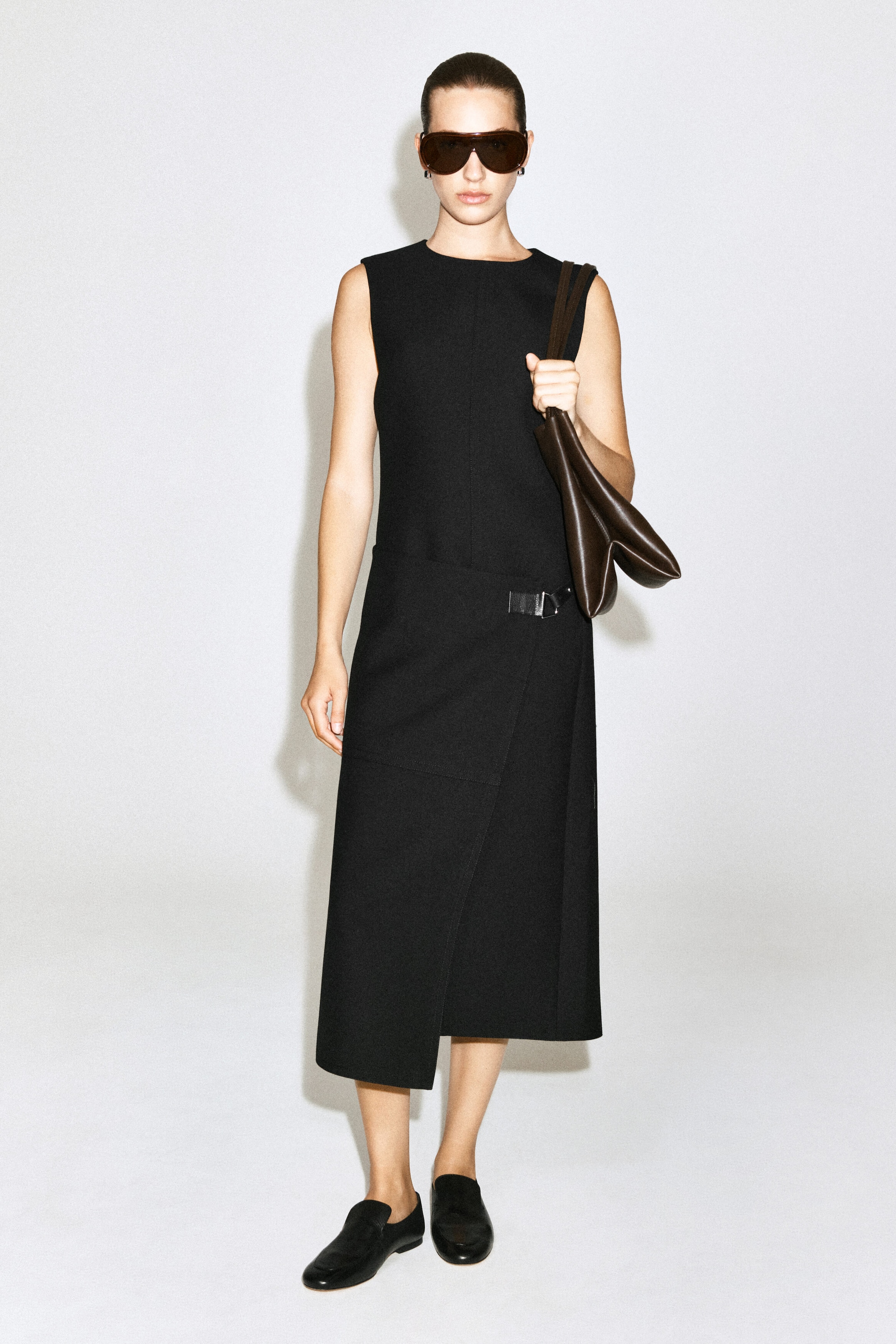 The model is wearing a black, belt-detail, double-faced wool midi dress featuring a sleeveless silhouette, a crew neck, and an asymmetrical wrap skirt with a silver buckle detail at the hip. The dress is styled with black loafers, dark sunglasses, and a brown leather shoulder bag carried by the model.