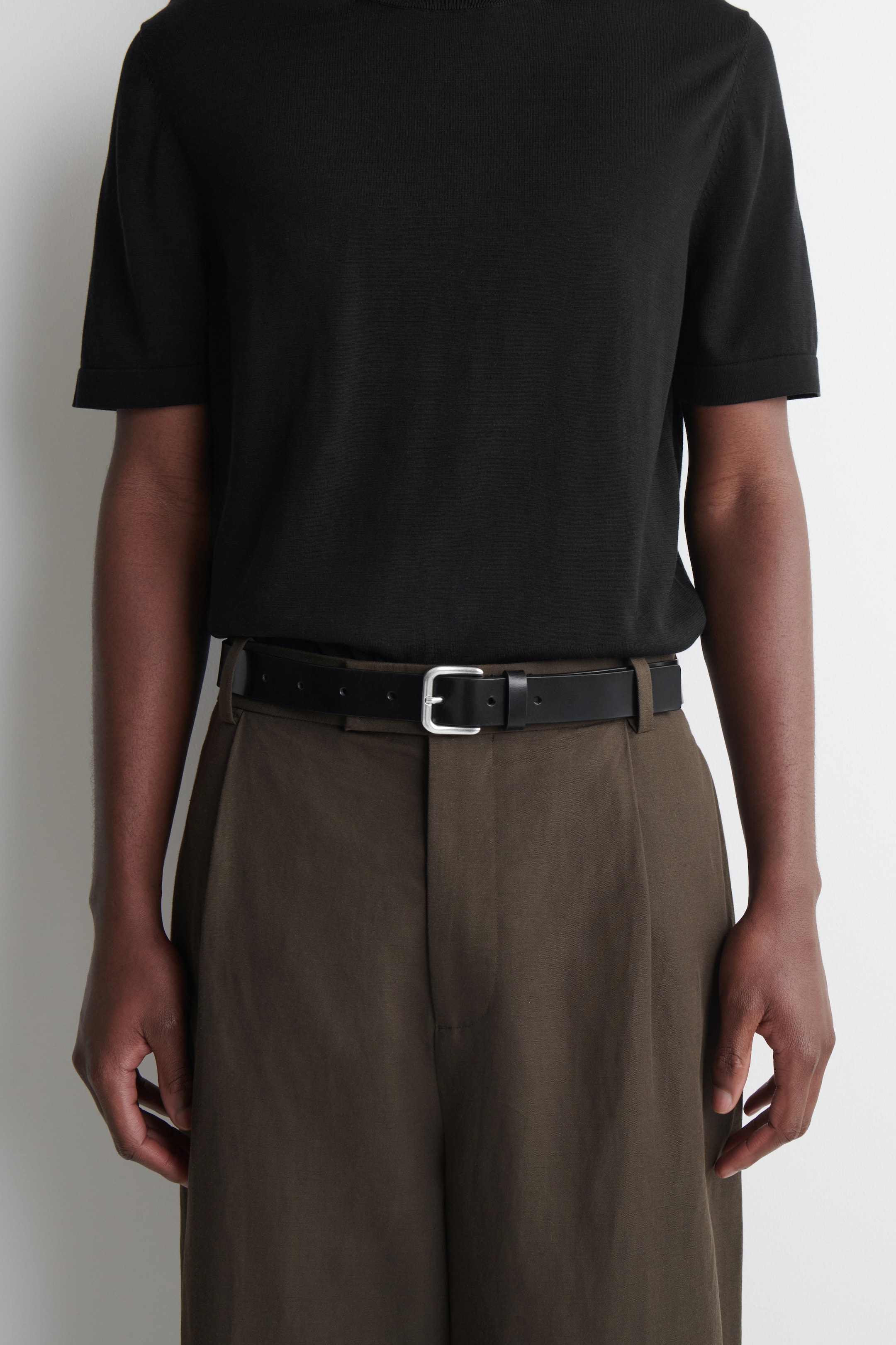 View larger image: A black leather belt with a silver-toned rectangular buckle fastens around the waist, securing brown trousers. The belt is visible from a front-facing angle, featuring a smooth, uniform strap.