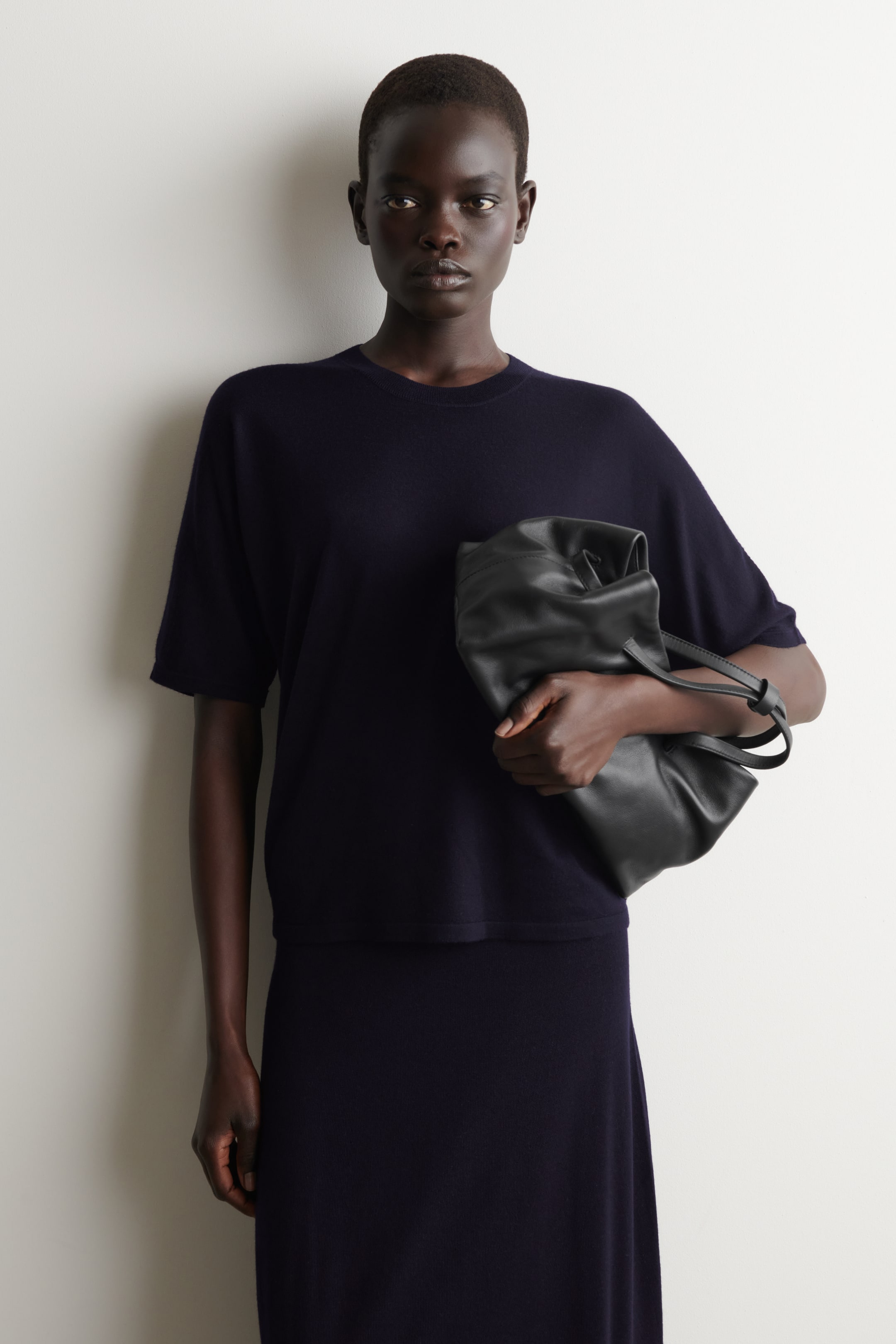 Grotere afbeelding bekijken: Een diepblauw gebreid t-shirt met korte mouwen en een geribbelde ronde hals wordt gedragen door het model, dat ook een bijpassende diepblauwe rok draagt en een zwarte tas vasthoudt.