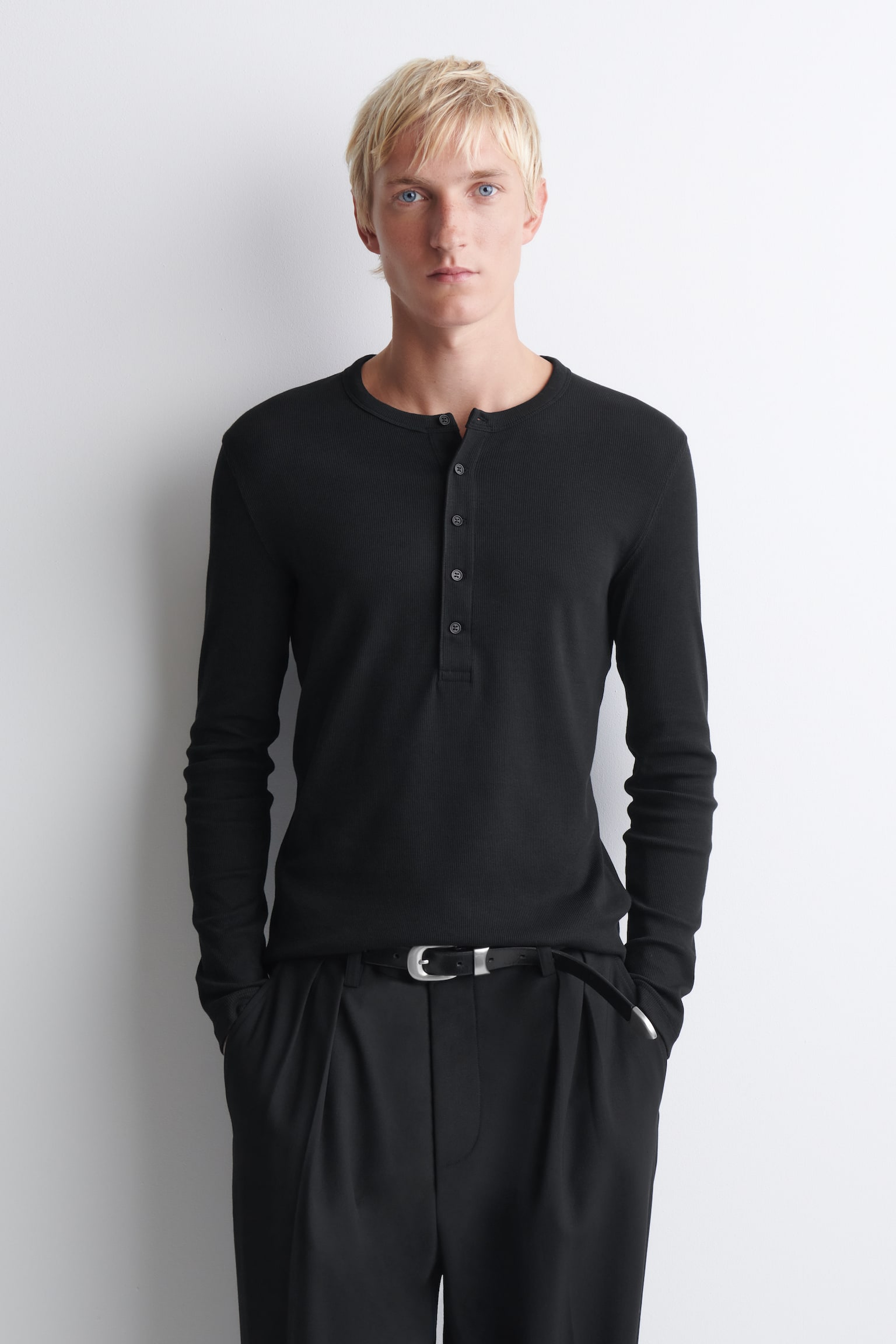 Das schwarze, schmale, gerippte Langarm-Henley-Shirt aus Baumwolle ist in der Vorderansicht zu sehen und zeigt seine körpernahe Silhouette sowie die feine Rippstruktur. Es verfügt über einen Rundhalsausschnitt mit geknöpfter Knopfleiste und wird in eine schwarze Hose mit Gürtel gesteckt getragen, um die klare Passform zu betonen. - 1