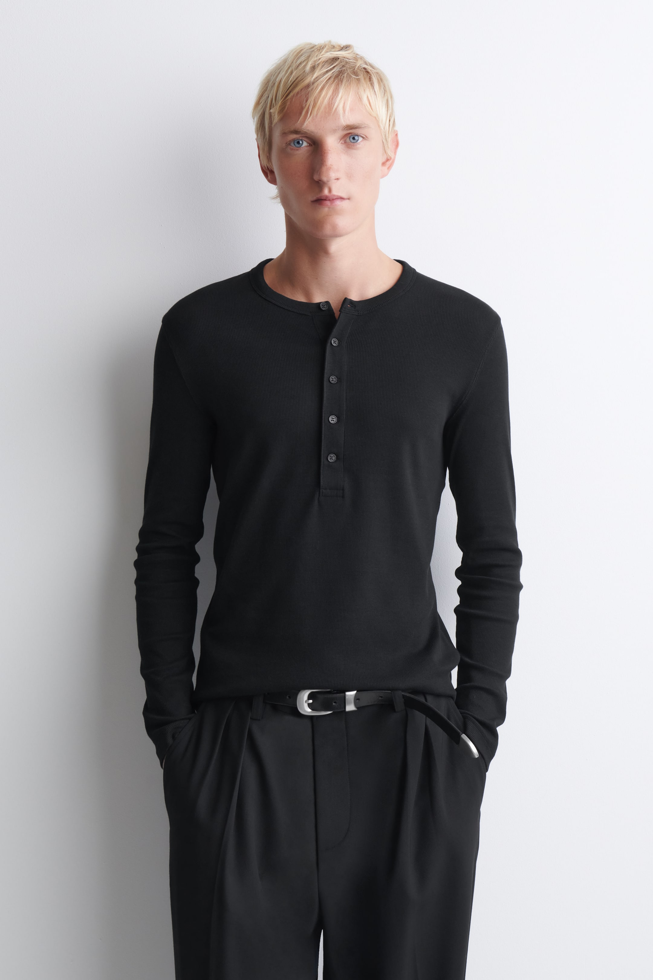 View larger image: The black slim ribbed cotton long-sleeved Henley T-shirt is shown from the front, showcasing its figure-hugging silhouette and fine ribbed texture. It features a round neckline with a button placket and is worn tucked into black trousers with a belt to highlight its neat fit.