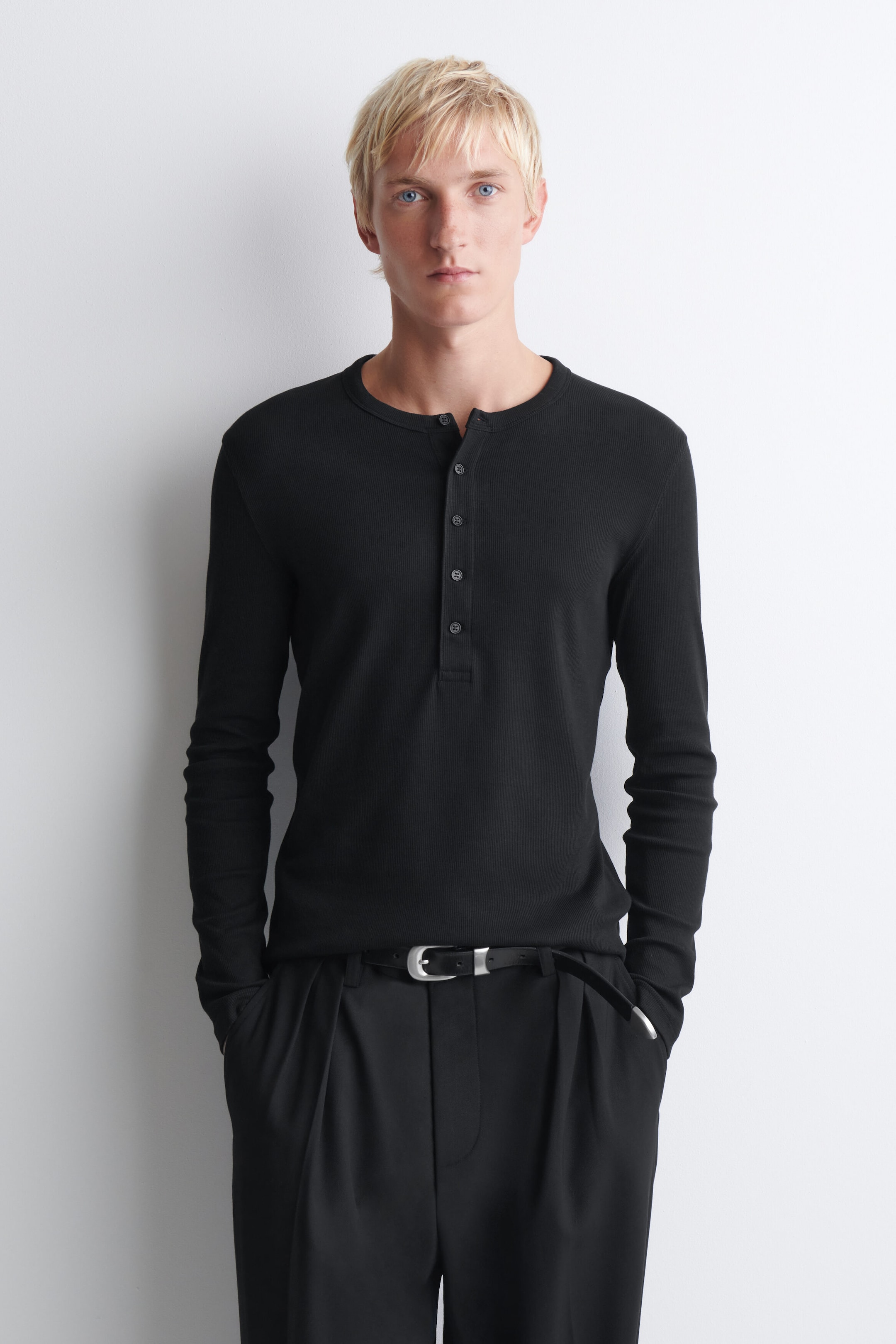 The black slim ribbed cotton long-sleeved henley T-shirt is shown from the front, highlighting its form-fitting silhouette and fine ribbed texture. It features a round neckline with a buttoned placket and is worn tucked into black trousers with a belt to emphasize its clean fit. The black slim ribbed cotton long-sleeved henley T-shirt is shown from the front, highlighting its form-fitting silhouette and fine ribbed texture. It features a round neckline with a buttoned placket and is worn tucked into black trousers with a belt to emphasize its clean fit.
