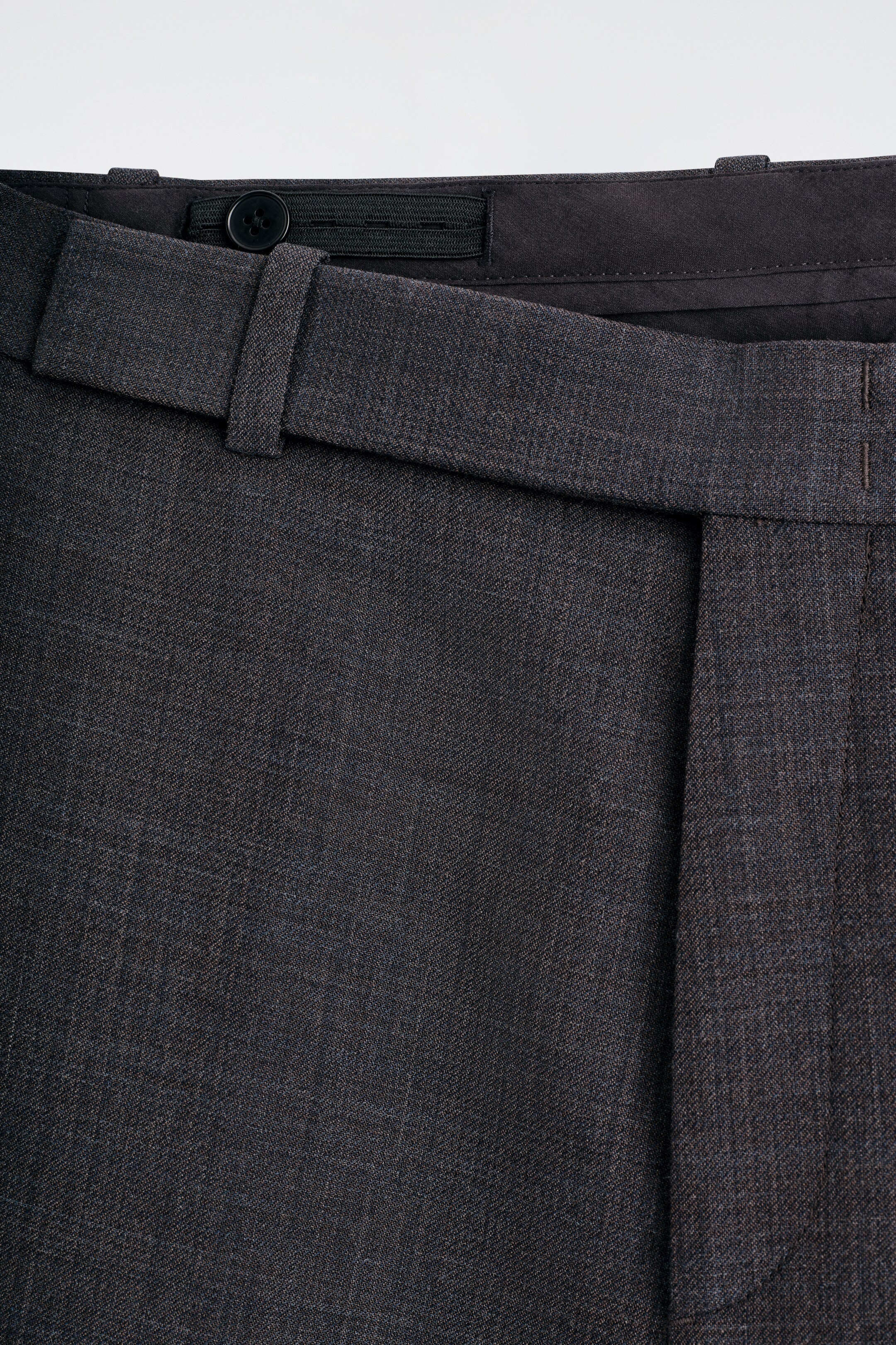 Vezi imaginea mai mare: Vedere din față a pantalonilor de culoare cărbune închis, cu un material texturat, o betelie vizibilă cu un nasture interior și închidere elastică, și un pliu vertical proeminent.