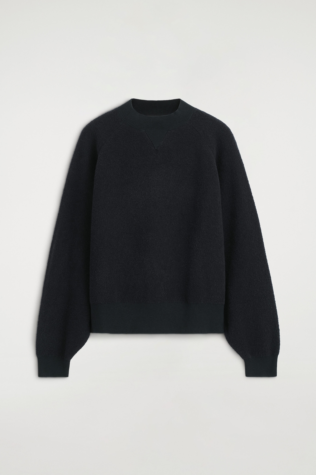A dark navy knit sweater is shown from the front, featuring a textured bouclé finish and a relaxed, sculptural silhouette. It is designed with a ribbed mock neck collar with a tonal V-insert, raglan sleeves, ribbed cuffs, and a ribbed hem.