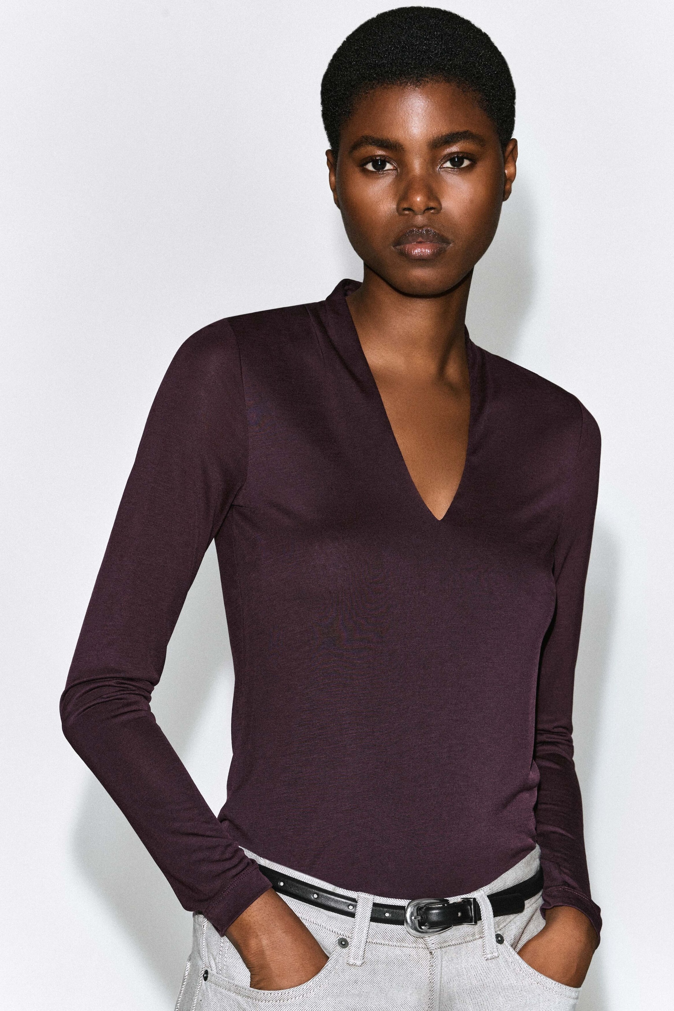 A dark purple, long-sleeved slim-fit top featuring a deep V-neckline with a draped collar is worn by the model. The top is styled tucked into light gray jeans with a black belt. A dark purple, long-sleeved slim-fit top featuring a deep V-neckline with a draped collar is worn by the model. The top is styled tucked into light gray jeans with a black belt.