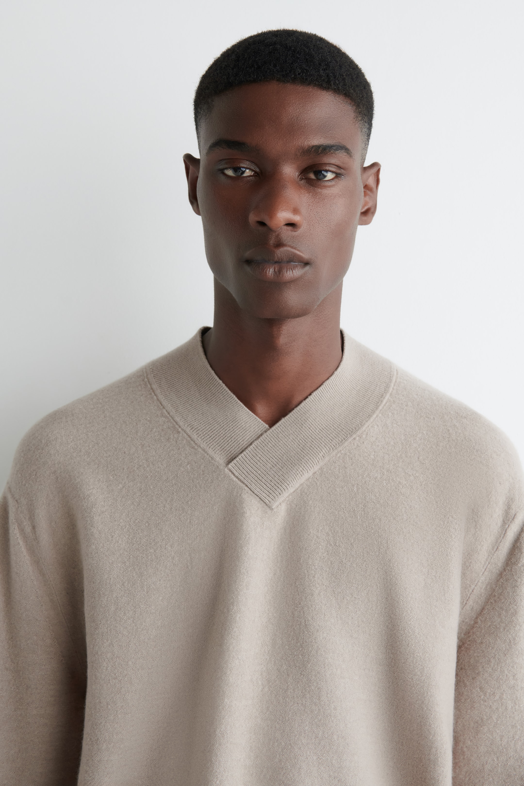 View larger image: A light beige V-neck sweater is being presented, featuring a wide, ribbed V-neck band. The sweater is exhibiting a soft, slightly brushed texture and a relaxed fit.