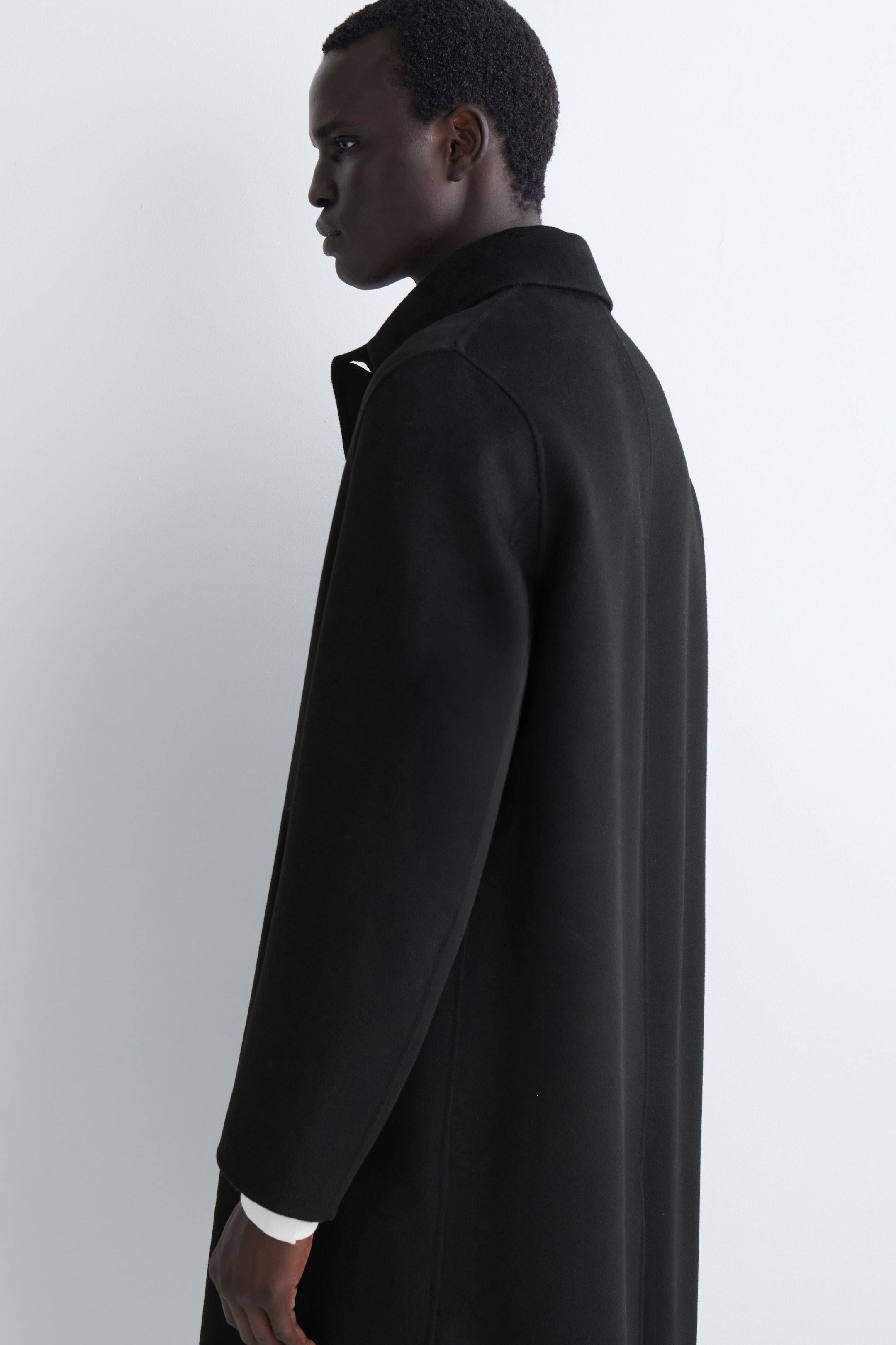 Visualizza immagine più grande: Un cappotto da auto di colore scuro viene presentato da un'angolazione laterale, mettendo in mostra la sua vestibilità rilassata e il colletto a punta. Le maniche lunghe coprono le braccia, con un polsino della camicia bianca che sbuca da sotto.