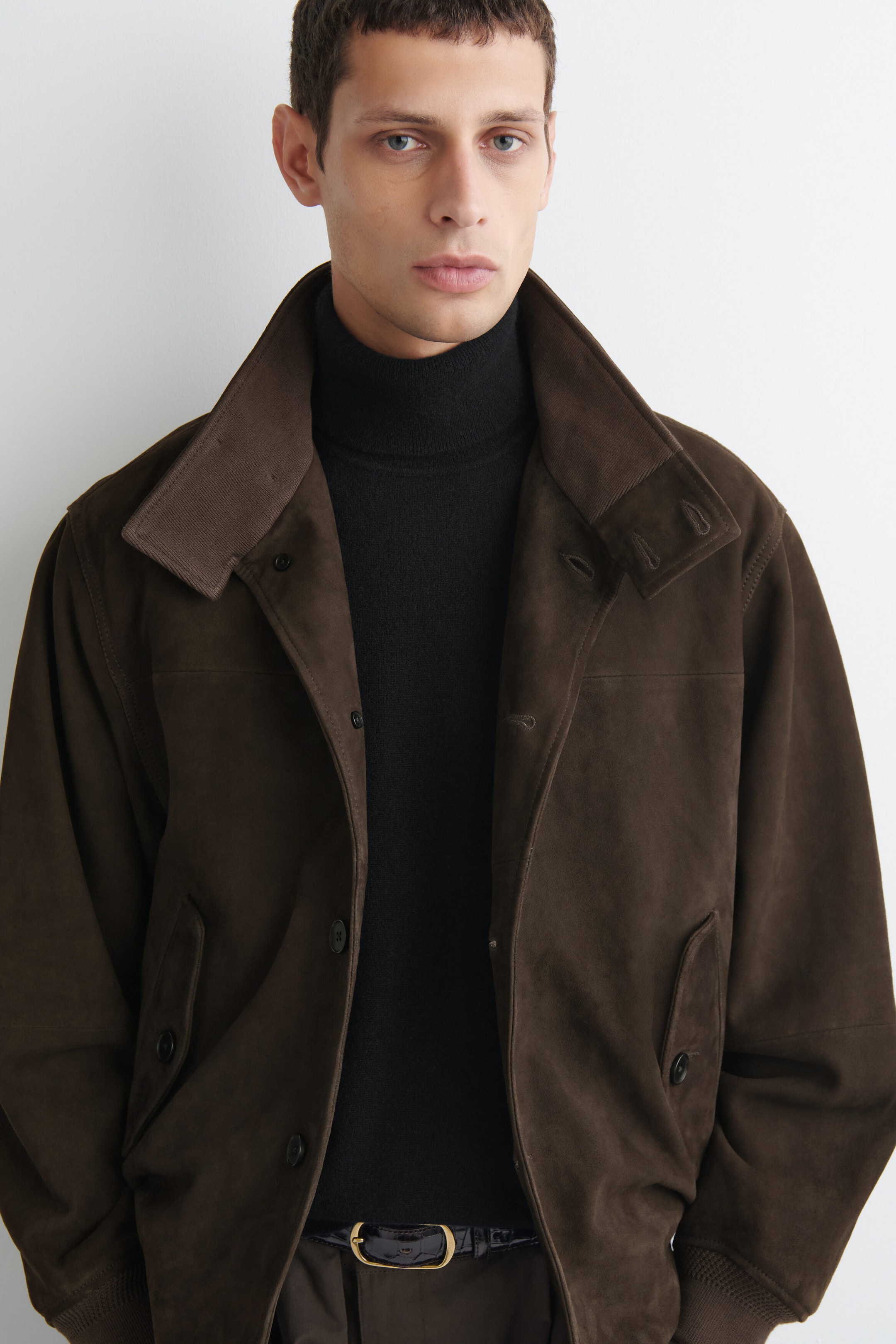 A brown suede jacket is shown from the front, featuring a wide collar with ribbed fabric trim, a button-front closure, and angled welt pockets. The jacket is layered over a black turtleneck sweater, highlighting its relaxed structure and ribbed knit cuffs. A brown suede jacket is shown from the front, featuring a wide collar with ribbed fabric trim, a button-front closure, and angled welt pockets. The jacket is layered over a black turtleneck sweater, highlighting its relaxed structure and ribbed knit cuffs.