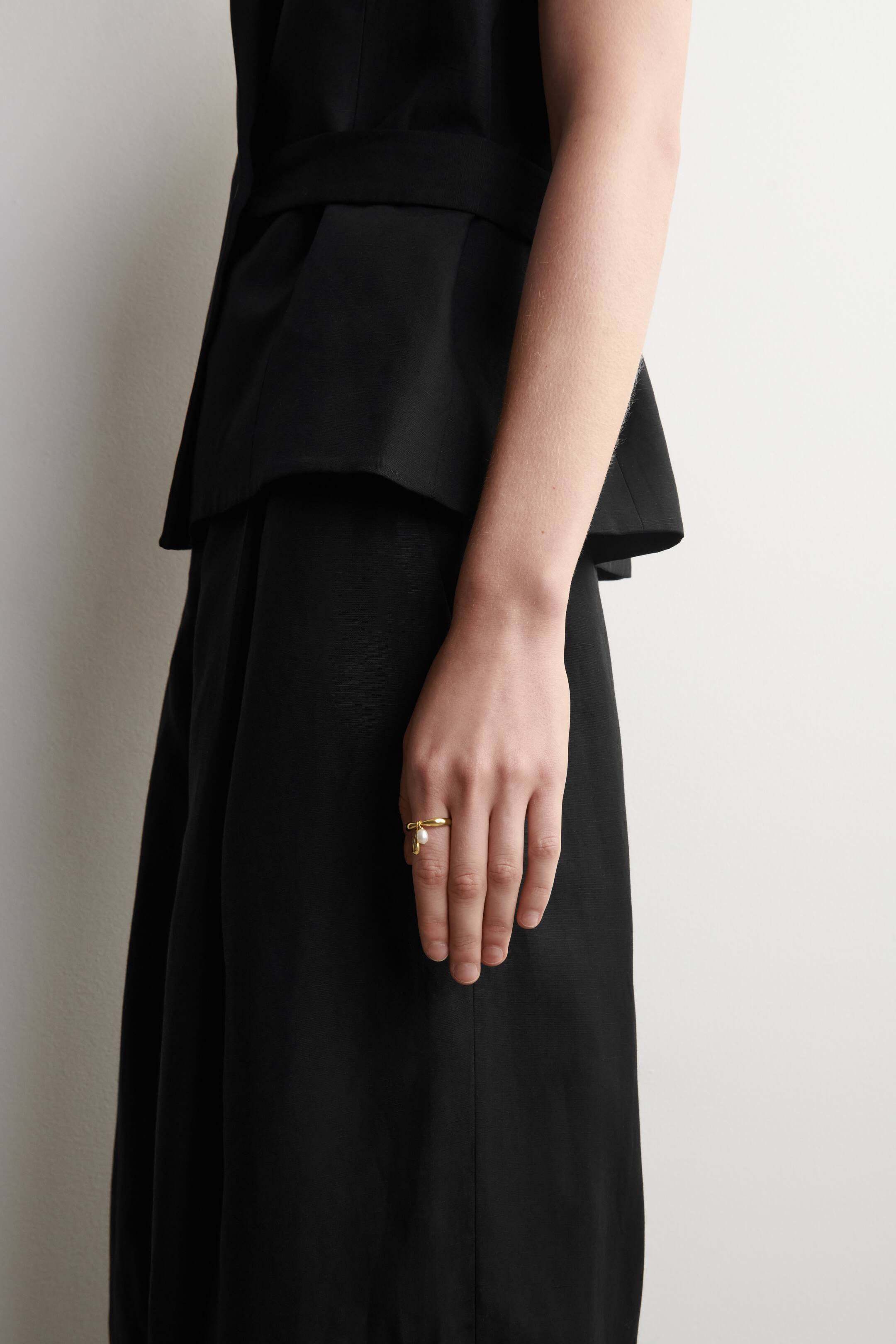 Grösseres Bild ansehen: Ein goldfarbener Ring wird an einem Finger getragen und zeigt ein einzigartiges Design, bei dem das Band sich selbst überkreuzt und eine tropfenförmige Perle trägt.